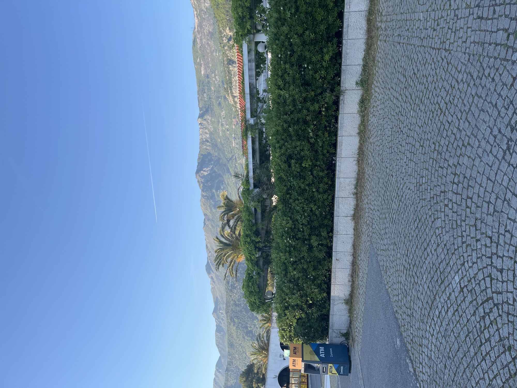 This image presents a scenic outdoor view, likely captured during the day in Budva, Montenegro, given the provided context. The frame is dominated by a vast, clear blue sky in the upper portion, through which a single white airplane contrail is visible.
In the background, majestic mountains rise, covered in green vegetation and rugged terrain under the bright sun. The mid-ground is characterized by dense, vibrant green foliage, including several palm trees and thick bushes, which seem to line a raised promenade or a building's perimeter. Peeking through the greenery and behind it, parts of a white building with arches and columns are discernible, hinting at a resort or hotel. Further up on the mountain slope, a distinctive long structure with multiple red-tiled roofs stands out.
The foreground features a pedestrian area paved with grey cobblestones, showing slight wear with sparse dry grass growing between the stones. This cobblestone path runs alongside a narrow stretch of asphalt road. On the lower left side, two ATM machines are placed, colored in blue and yellow. Visible text on these machines clearly reads "ATM", "Euronet", and "Cash 24/7". The scene is tranquil, devoid of people, and appears to be under clear, sunny weather, likely in the morning or late afternoon.