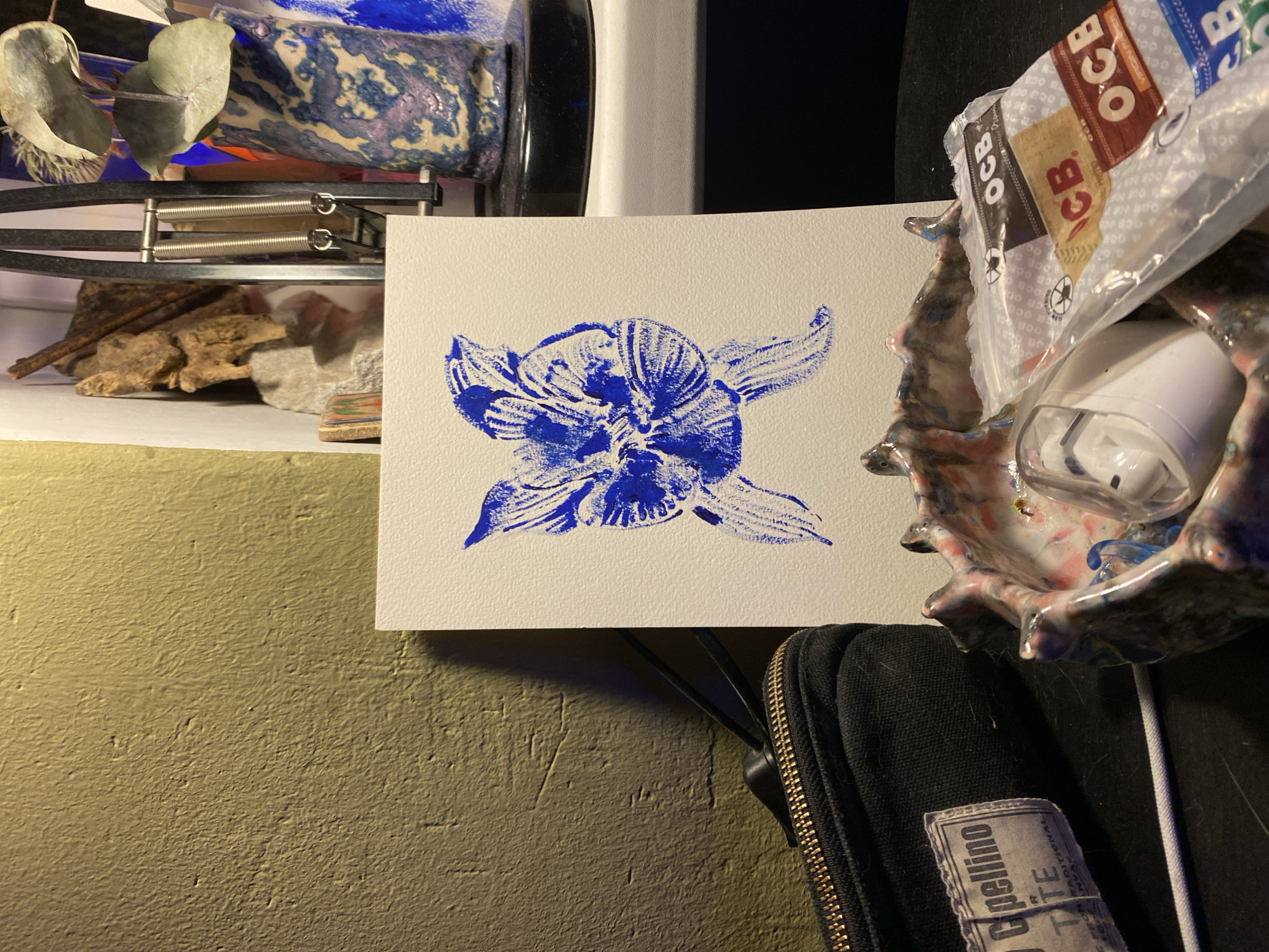 This image presents a close-up view of a personal space, likely a shelf or desk in a room in Moscow, Russia, illuminated by indoor artificial lighting. The central focus is a white piece of paper bearing a vibrant blue monoprint or stamp. The print depicts a stylized figure with prominent ear-like extensions, strongly resembling Grogu (Baby Yoda) from Star Wars, though it could also be interpreted as a unique floral design.
To the left of the print, an assortment of natural and functional items is arranged. These include dried eucalyptus leaves, pieces of natural driftwood or bark, and several rocks. Above these objects, a black metal articulated arm with visible springs, characteristic of a desk lamp or microphone stand, is partially seen. Further back, a blue and purple swirled ceramic or glass object is visible.
On the right side of the frame, adjacent to the print, an irregularly shaped ceramic dish with a multi-colored glaze (featuring tones of pink, blue, and white) holds a white AirPods charging case. Above this, a clear plastic bag contains multiple packs of "OCB" rolling papers. Visible text on the packaging includes "OCB" (repeated), "Hermp" (likely 'Hemp'), "VIRGIN PAPE" (part of 'Virgin Paper'), and numbers such as "20500" and "300 P".
Beneath the central print and extending towards the bottom right, a black zippered pouch or case is visible. It features a worn label with partially decipherable text including "Opellino," "TITE," "FRON RADITIONAL" (likely 'FROM TRADITIONAL'), and "INVAR." The background throughout the image is a textured, olive-green painted wall. The overall scene suggests a personalized and somewhat eclectic environment.