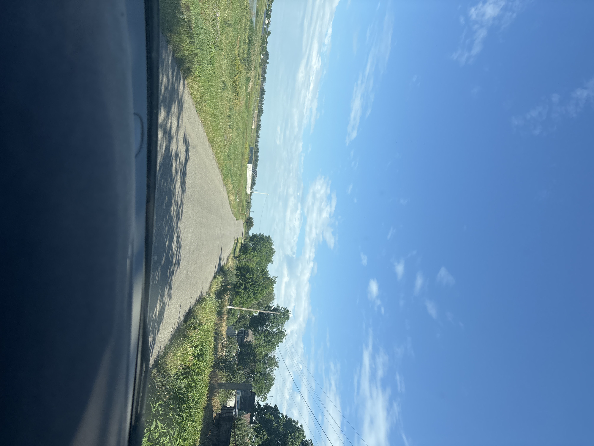 The image depicts a rural road scene under a bright blue sky with scattered white clouds. The perspective is from inside a vehicle, showing the dashboard in the foreground. The road, paved with asphalt, stretches into the distance, lined by lush green grass and vegetation on either side. Trees and houses are visible on the right side of the road, suggesting a small settlement. On the left, a body of water and more vegetation can be seen. Utility poles with power lines are also present, stretching along the right side of the road. The overall impression is of a sunny day in a quiet, possibly agricultural, area.