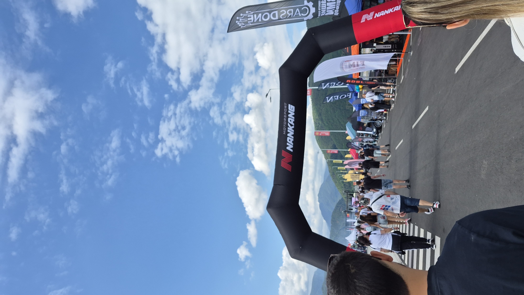 In an outdoor scene, a black inflatable archway with "N NANKANG MOTORSPORT" emblazoned on it stands across a paved road. The archway is set against a backdrop of rolling hills covered in green foliage under a partly cloudy blue sky. A crowd of people walks along the road, appearing to be at an outdoor event or exhibition. Various tents and flags are visible on either side of the road, suggesting a marketplace or festival setup. In the foreground, the backs of two individuals' heads are visible, one with dark hair and sunglasses, and another with blonde hair. The overall atmosphere is lively and suggests a public gathering or event.
