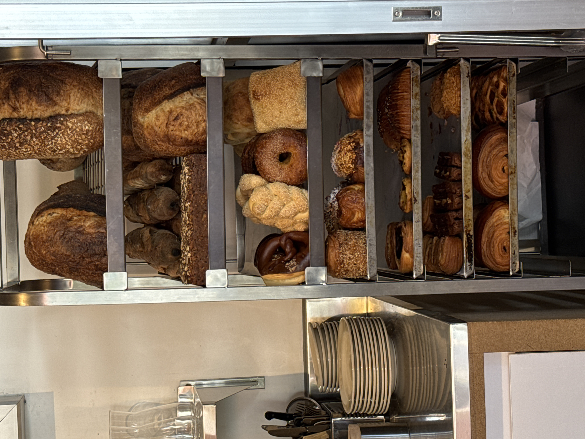 The image shows a display case of baked goods, likely in a bakery or cafe. The case is made of metal and has multiple compartments. The baked goods include various types of bread, bagels, croissants, and pastries. The plates and cooking utensils beneath the bakery products give a hint of the operation of the bakery. The location is in the city of North Kuta, Indonesia.