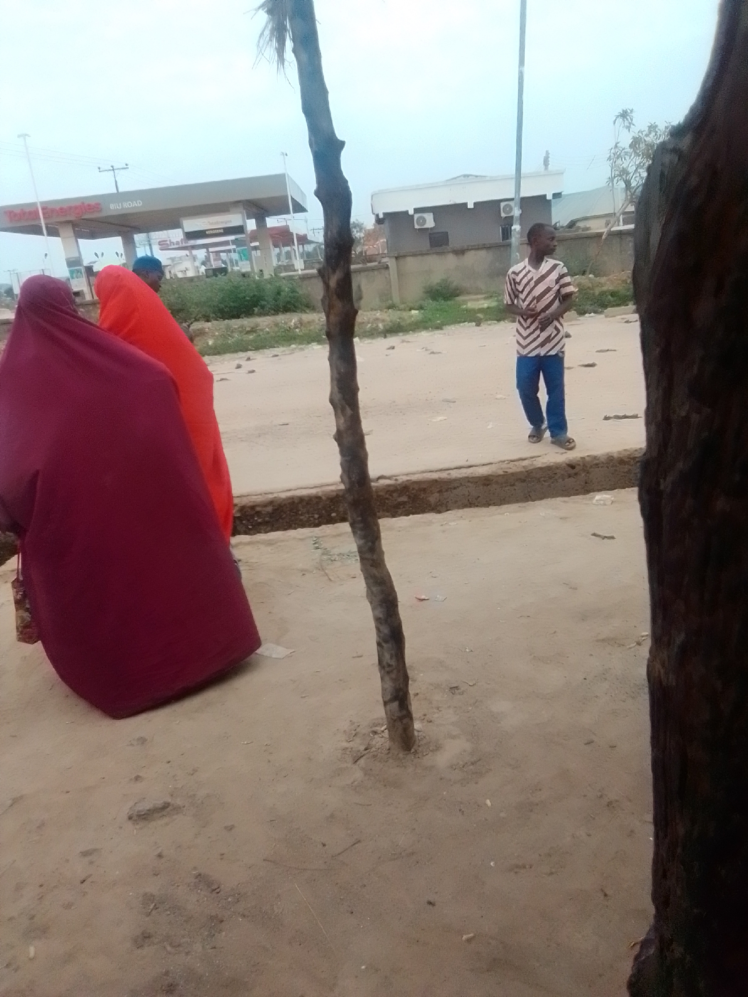 In Gombe, Nigeria, a man in a striped shirt and blue pants stands on a dusty road. To his left, two women are partially visible, one in a maroon hijab and another in an orange hijab. In the background, a Total Energies gas station with its distinctive branding is present, along with the "BIU ROAD" sign. The environment appears to be an outdoor, somewhat undeveloped area with a cloudy sky. A prominent tree trunk stands in the foreground, obscuring a portion of the scene.