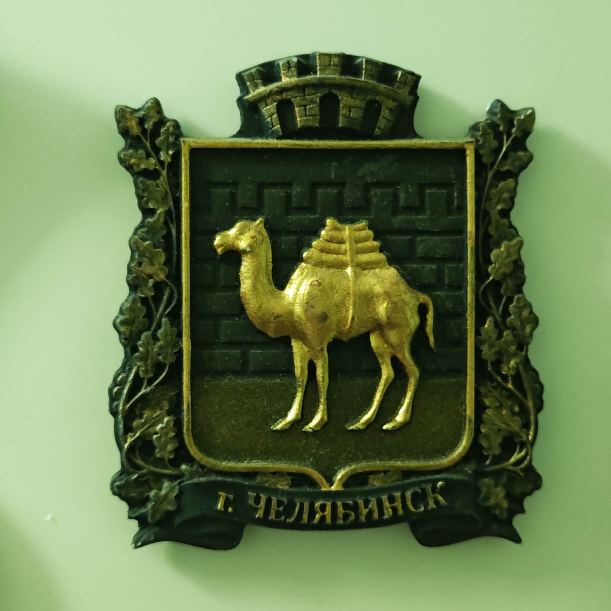 This media file captures a detailed image of a heraldic emblem, likely a municipal coat of arms, mounted on a light green wall. The emblem is shield-shaped, rendered in a metallic finish, predominantly gold and dark green or black.
The central feature of the emblem is a golden, two-humped camel (Bactrian camel), depicted standing and seemingly laden with goods or equipment. This camel is set against a dark background textured to resemble a brick wall. Above the main shield, a golden mural crown, a common symbol for cities or fortified settlements, is displayed. The sides of the shield are adorned with intricate, dark-colored foliage, possibly oak leaves and branches, providing an ornamental border.
At the bottom of the emblem, a dark banner unfurls, bearing golden text in Cyrillic. The visible text reads "Г. ЧЕЛЯБИНСК". This translates to "City of Chelyabinsk," identifying the emblem as the coat of arms for the Russian city of Chelyabinsk. The camel on the coat of arms traditionally symbolizes the city's historical role as a trade hub.
The object itself appears to be a physical plaque or relief, possibly made of metal such as brass or bronze, given its texture, sheen, and the way light reflects off its surfaces. It is displayed indoors, with soft lighting creating subtle shadows on the wall behind it. No specific activity or event is taking place; the image is a static portrayal of the emblem.