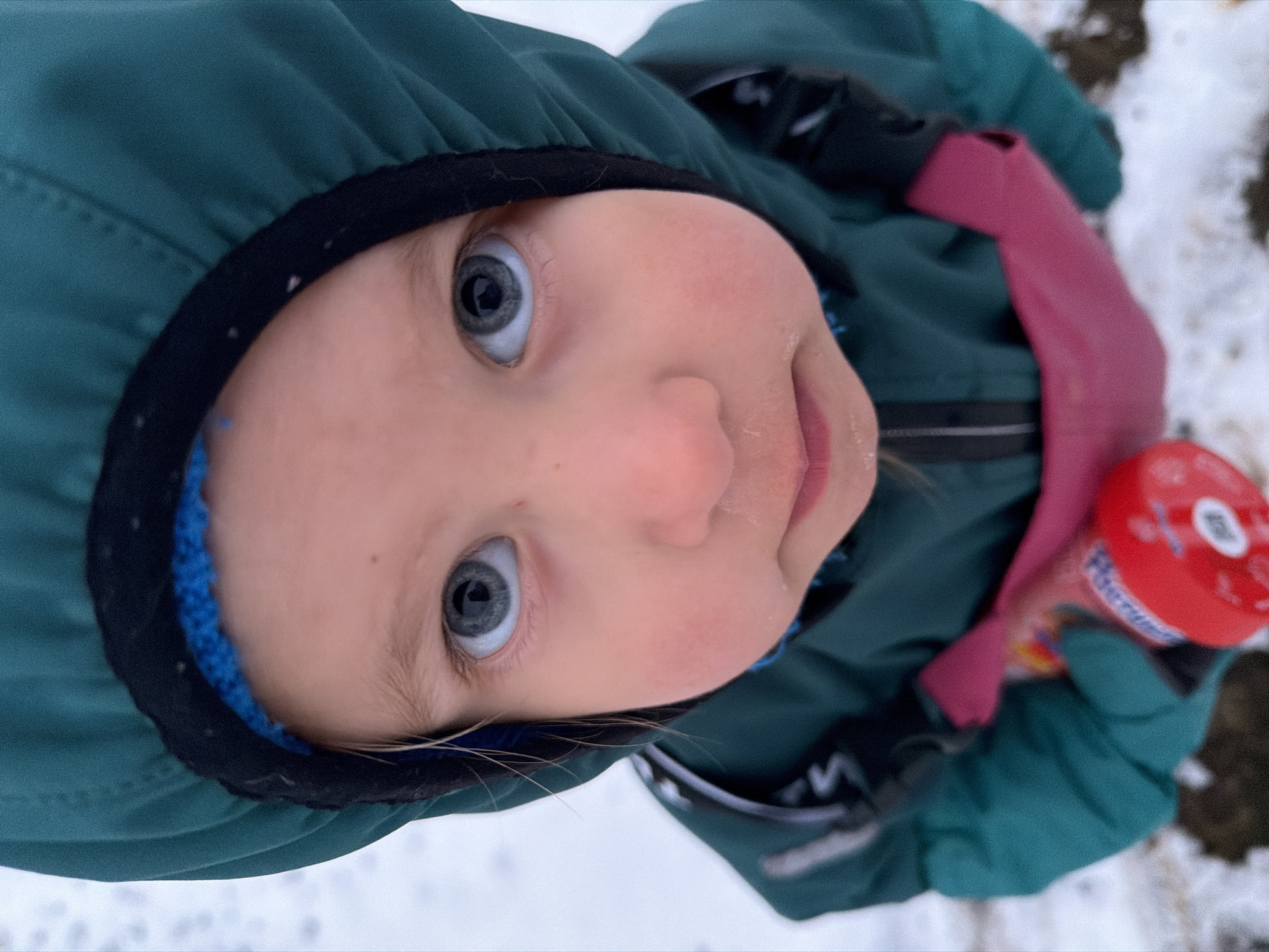 A young child with fair skin and light blue eyes is the central focus of the image. The child is looking upward at the camera, wearing a green hooded jacket with a black trim. A blue knitted material lines the inside of the hood. The child is also wearing pink gloves and holding a red cylindrical object in their gloved hand. The background is a mix of white, likely snow, and darker patches, possibly grass or dirt. The picture was taken outdoors. It is not possible to confirm the location of the city of Chaikovsky, Russia, from the visual data provided.