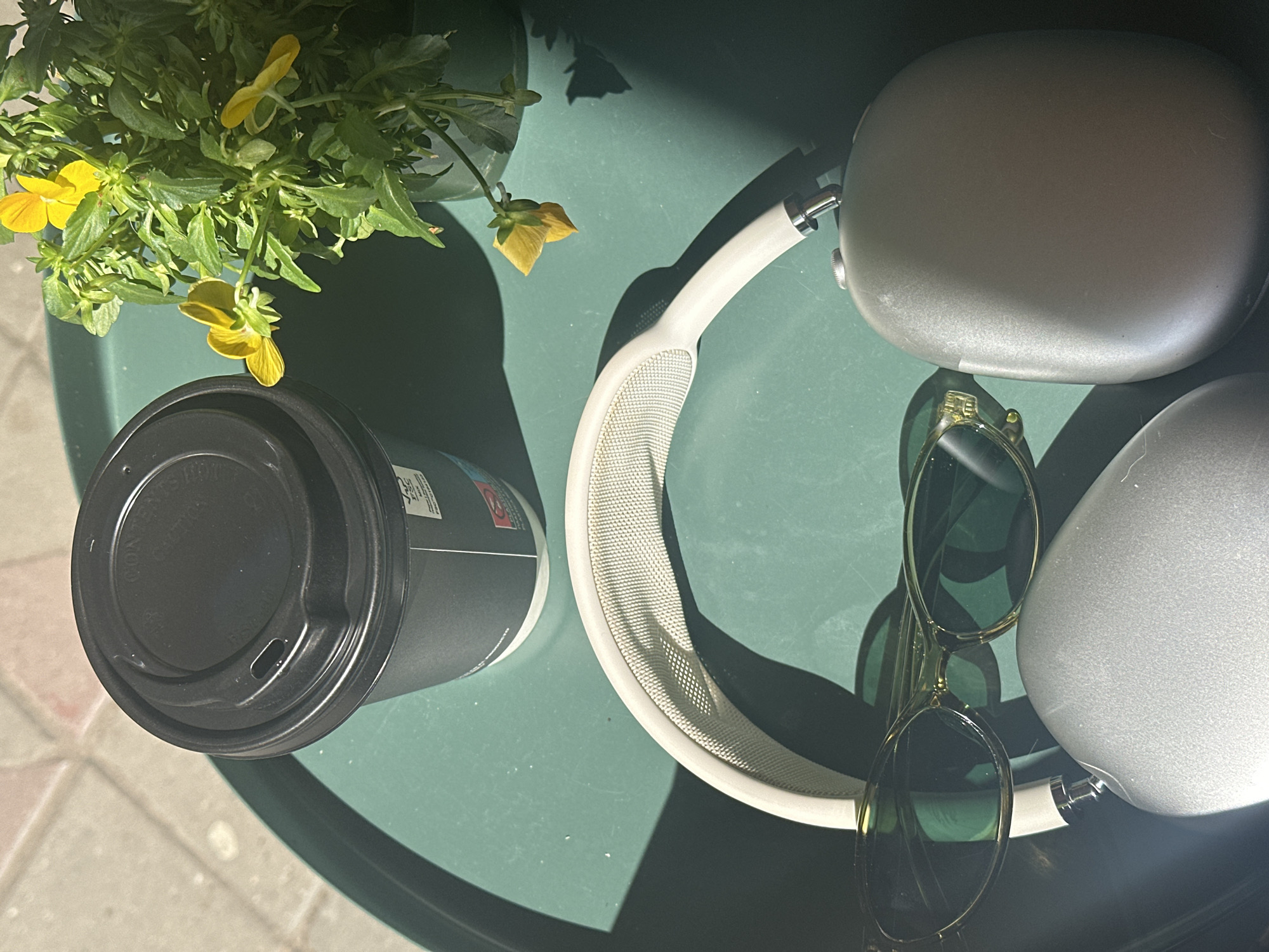 This overhead image depicts a relaxed outdoor scene on a bright, sunny day in Novi Sad, Serbia. Various personal items are arranged on a dark green, round outdoor table, creating a sense of leisure.
In the upper left, a potted plant with lush green leaves and several vibrant yellow flowers, resembling violas or pansies, adds a touch of nature to the setting. Its dark pot is partially visible. Strong, defined shadows are cast by the plant and other objects, indicating direct sunlight, likely during late morning or early afternoon.
Centrally placed are a pair of white or light grey over-ear headphones, identifiable as Apple AirPods Max by their distinctive mesh headband and ear cup design. One ear cup, made of a metallic material, reflects the light. Next to them, a pair of stylish sunglasses with light-colored frames and dark green lenses rests on the table.
To the left of the headphones, a black disposable coffee cup with a matching black plastic lid is positioned. The lid shows faint, unreadable embossed text. Some indistinct branding or text is also visible on the side of the cup.
The table surface appears smooth and slightly reflective. In the bottom left corner, part of the ground is visible, consisting of light-colored paving stones or tiles, reinforcing the outdoor setting. No people are present in the image, but the arrangement suggests someone has momentarily set down their belongings while enjoying a break.