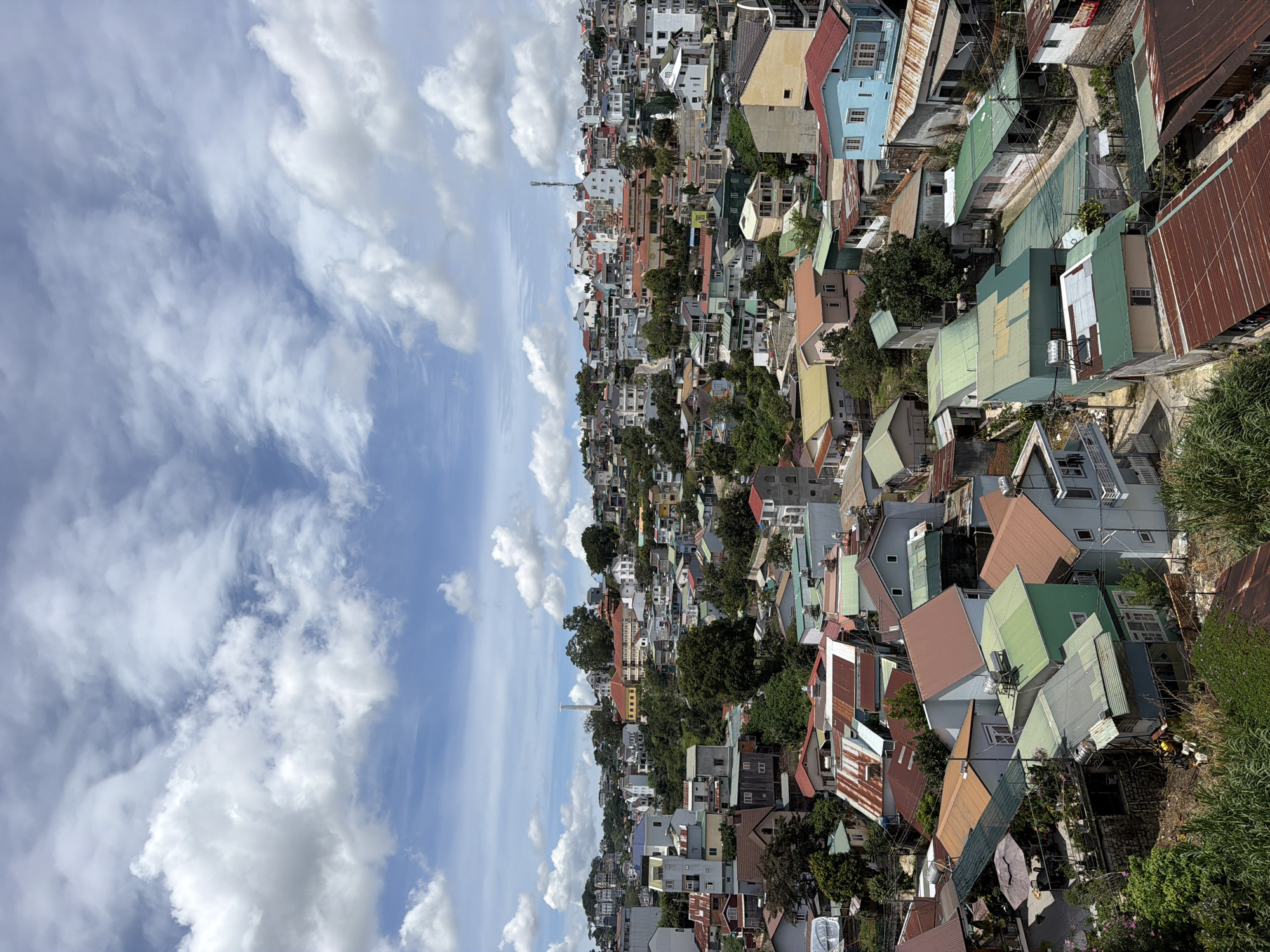 This is an aerial shot of a dense urban landscape in Da Lat, Vietnam, on a cloudy day. The image is dominated by a multitude of houses with various colored roofs, including red, green, blue, and brown, clustered together on a hilly terrain. Some houses feature corrugated metal roofs, indicating a mix of housing styles. Amidst the buildings, there are patches of lush green trees and vegetation, suggesting the city's verdant environment. In the distance, a prominent tower and a statue are visible, along with a larger, yellow-colored building complex. The sky is filled with dramatic cumulus clouds against a bright blue backdrop. No people are visible in the frame, and the scene appears to be a quiet, residential area.