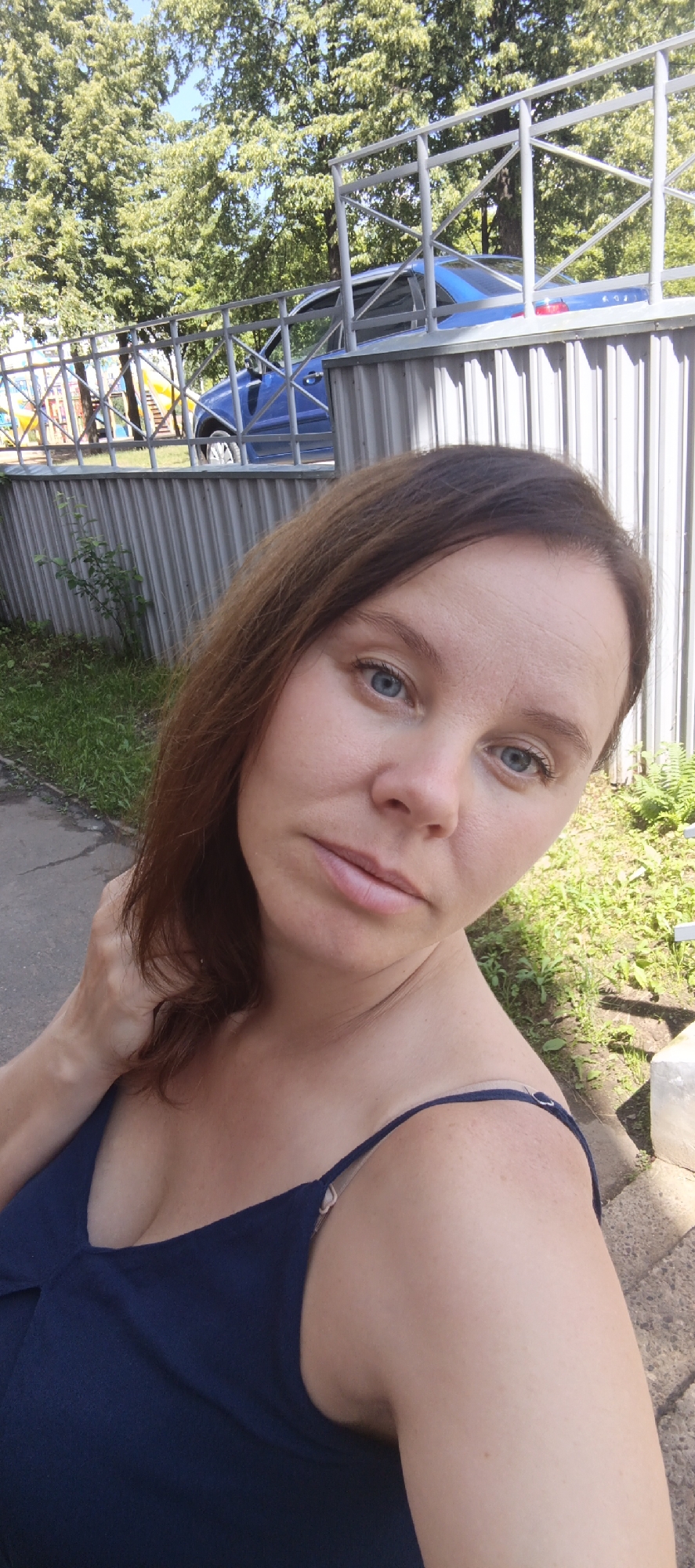 The media file is a selfie taken outdoors in Chaikovsky, Russia. The subject is a woman with brown hair and blue eyes, looking directly at the camera with a neutral expression. She is wearing a dark blue camisole. The background shows a metal fence with a blue car visible behind it, and lush green trees and foliage. The scene suggests a sunny day, possibly in the late morning or early afternoon, given the bright lighting. A paved pathway is visible in the lower left portion of the image, and some grass and greenery on the right. There is no visible text in the image. The overall impression is a casual, outdoor portrait.