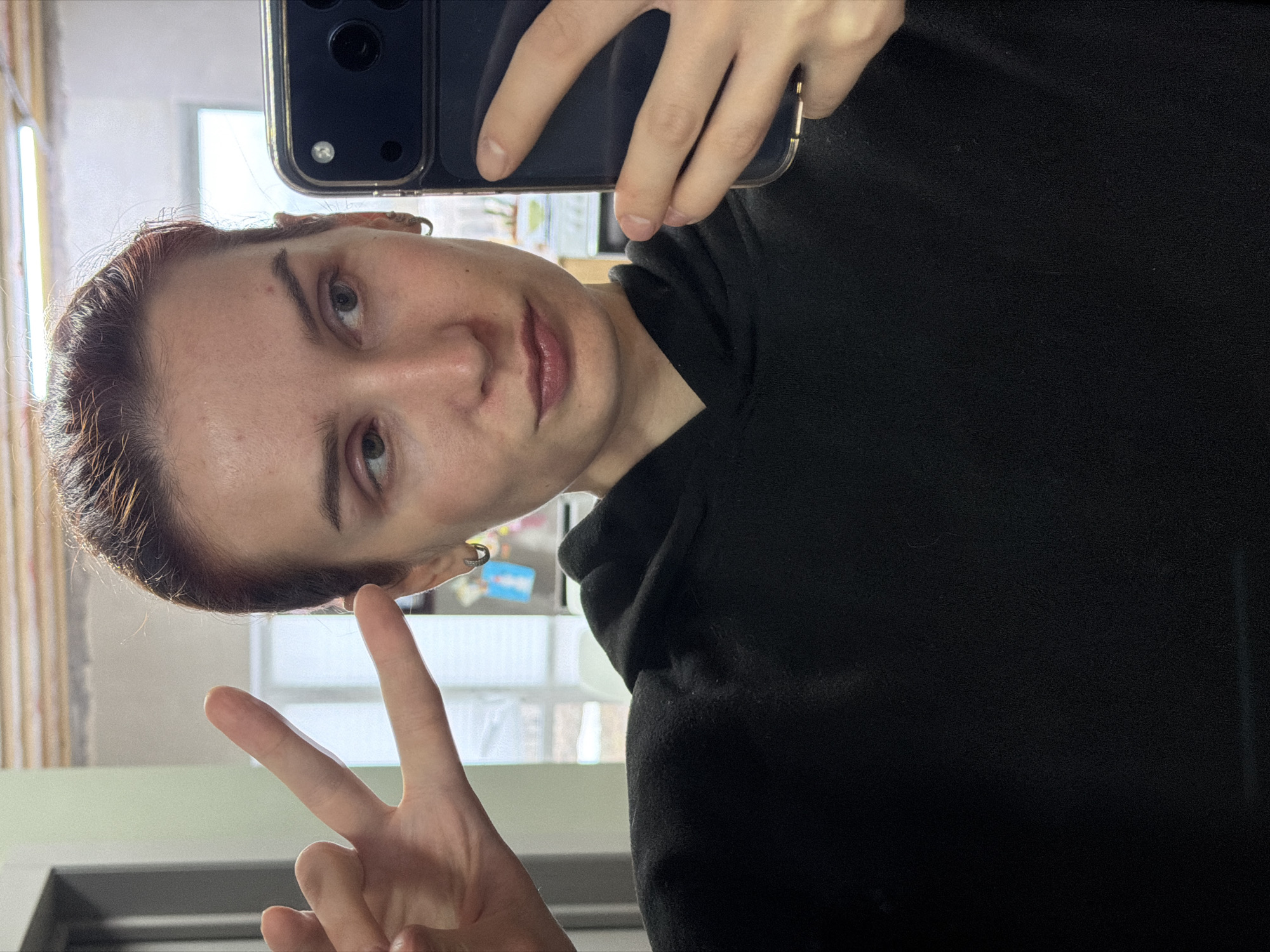 A person with short, reddish-brown hair is taking a selfie while making a peace sign. They are wearing a black hooded sweatshirt. The phone used for the selfie is a dark color with multiple camera lenses. The background appears to be a room with a window and blinds. Based on visual information, there is nothing to indicate that the image was taken in Chaikovsky, Russia.
