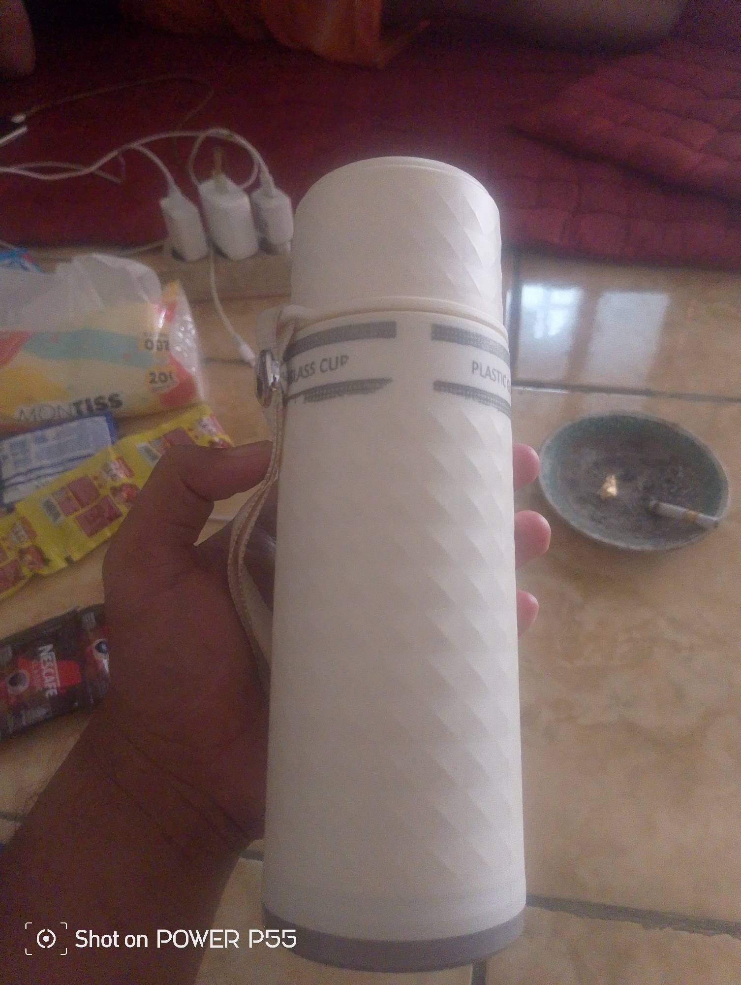 In Lebaksiu, Indonesia, a person holds a white cylindrical thermos with a textured surface and a beige strap. The thermos has two gray bands, one with the text "GLASS CUP" and the other with "PLASTIC." In the background, a power strip with three white adapters is visible on a red surface. To the left of the thermos, there are several packets, including one labeled "MONTISS" and a red packet with "NESCAFE" printed on it. To the right of the thermos is a small ashtray containing a lit cigarette. The scene appears to be indoors, possibly in a residential setting.