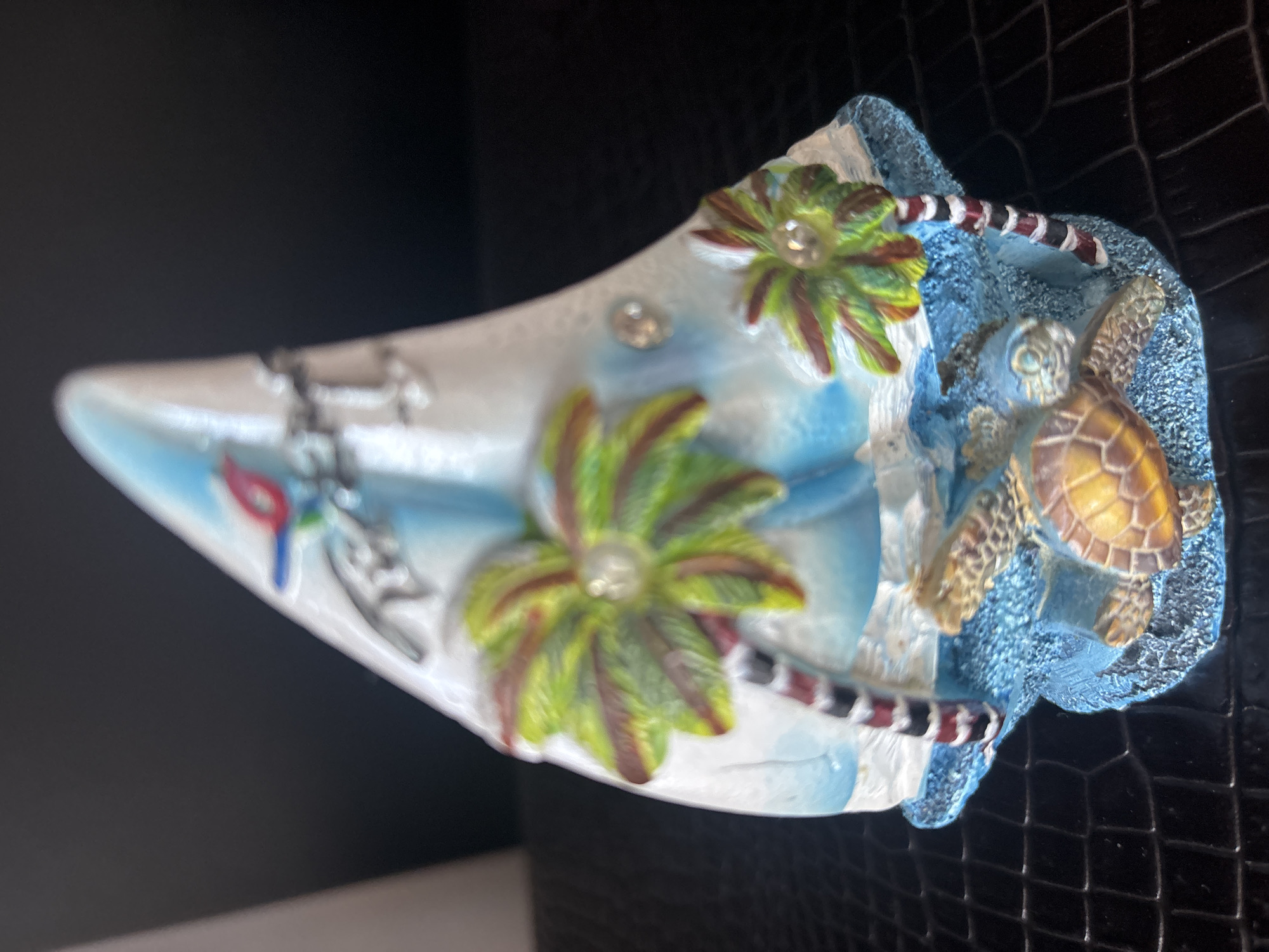 This is a close-up, eye-level, full-body shot of a decorative ceramic figurine on a dark, textured surface, possibly a table or shelf. The figurine is shaped like a sailboat with a white sail that has blue wave-like accents. It is adorned with several decorative elements: two palm trees with green fronds and red and black striped trunks, two sea turtles with yellow and brown shells, and a small red and blue bird perched near the top of the sail. Clear, faceted embellishments, possibly rhinestones, are placed at the center of each palm frond and on the sail. The base of the figurine is blue and textured to resemble the ocean. The background is dark and out of focus. The location in Dubai, United Arab Emirates, is not discernible from the image.