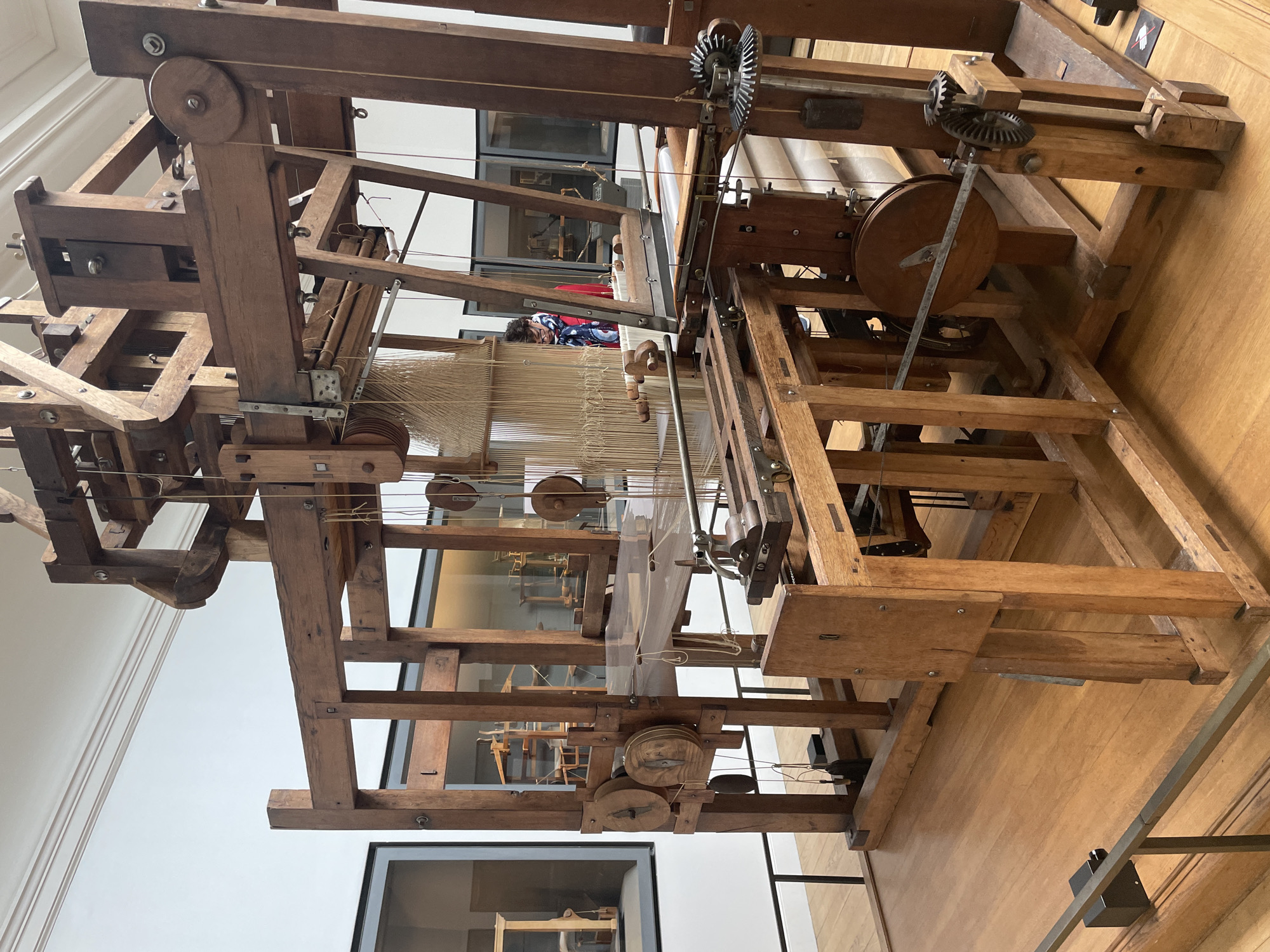An imposing, antique wooden loom stands in a well-lit museum hall with polished wooden floors. The loom is intricate, featuring numerous wooden beams, spools, pulleys, and threads. A person with dark hair, wearing glasses and a red jacket with a colorful scarf, is visible behind the loom, observing it. In the background, through glass displays, other historical looms are visible, suggesting the setting is a museum dedicated to textile machinery or industrial history. The overall impression is of a meticulously preserved piece of engineering from a bygone era. The location of this image is in Paris, France.