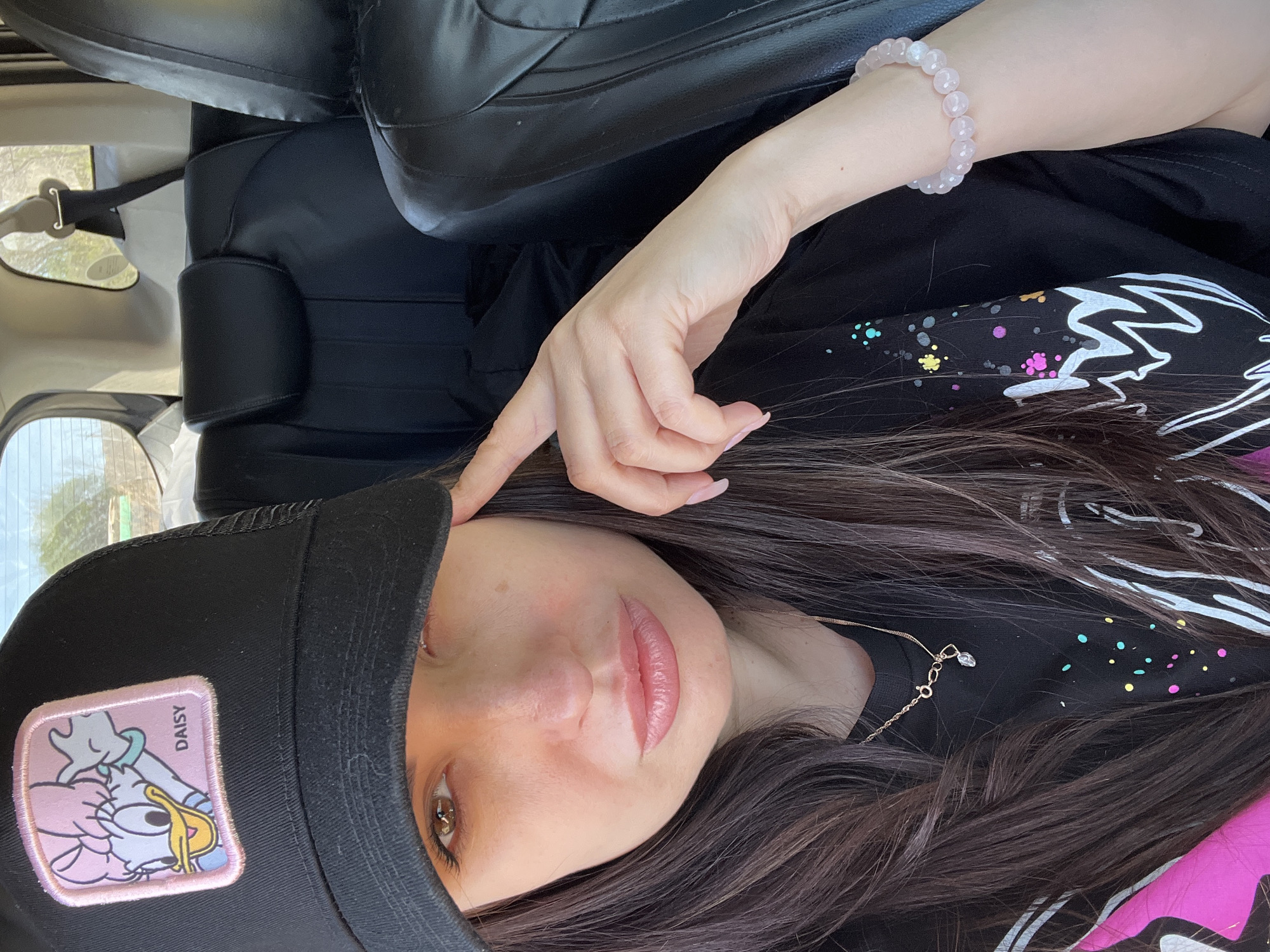 A selfie shot of a woman in a vehicle. She is wearing a black baseball cap with a Daisy Duck patch on the front. She has long brown hair, light skin, and is wearing a necklace. Her right hand is near her face. She is also wearing a pink beaded bracelet on her right wrist. The vehicle has black leather seats. The location appears to be outdoors, possibly in a vehicle traveling in an unknown city in Unknown.