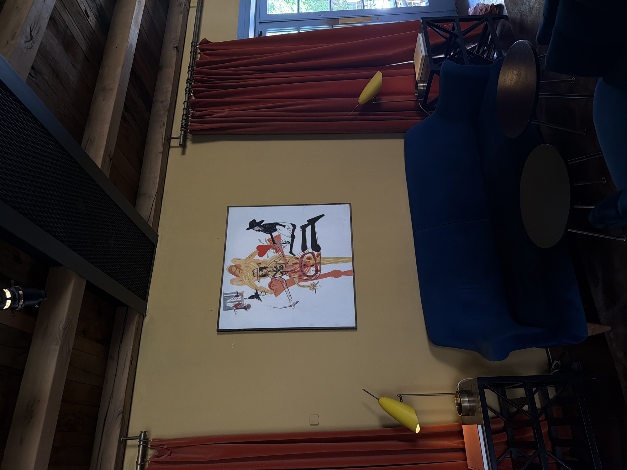 The image depicts an interior scene, likely a room with a rustic and modern aesthetic. Prominently featured is a large, dark blue sofa or lounge chair, partially draped with fabric. Beside the sofa, two round, metallic side tables are visible. A framed artwork hangs on a tan-colored wall, showcasing a surreal illustration of figures, possibly a stylized depiction of anatomical elements and human forms. The artwork has a white background and appears to be a print or painting.
Above the artwork, a section of dark wood beams and a black mesh-like structure are visible, suggesting a high ceiling or loft-style architecture. A red velvet curtain is drawn across a window or opening, creating a dramatic visual element. Another red curtain is visible lower down on the wall. A yellow, pear-shaped lamp extends from a metal rod, casting a warm light.
In the foreground, partially obscured by the sofa, is a black metal structure that could be a shelf or part of a piece of furniture. A black spotlight fixture is mounted on a wooden beam on the left side of the image. The overall impression is of a stylish and perhaps artistic space, with a blend of natural materials and contemporary design. The lighting suggests it could be evening or an interior space with controlled lighting. No visible text is present in the image. The location context provided is Tbilisi, Georgia.