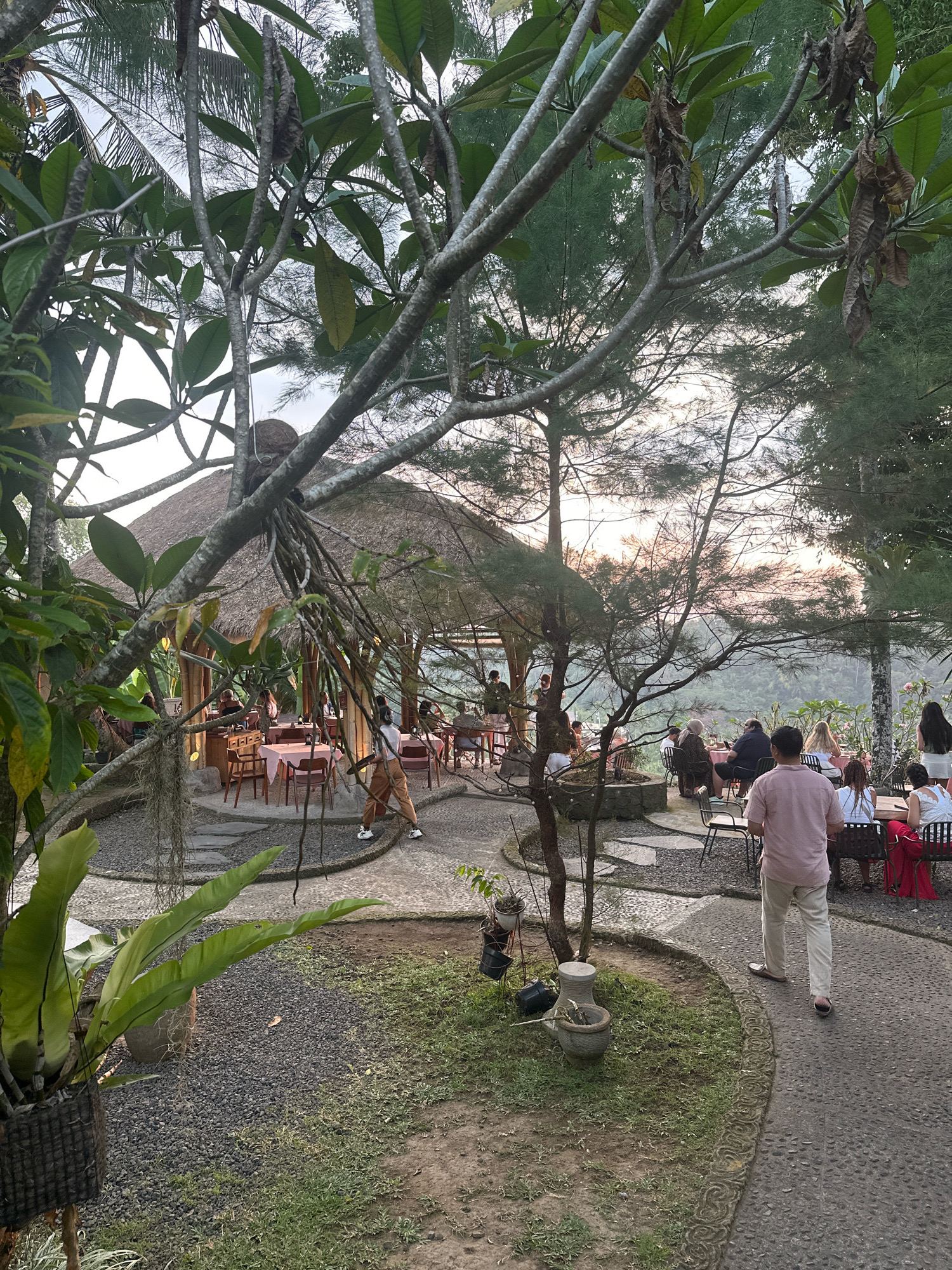 An outdoor dining area with a thatched-roof building and tables set for guests is nestled among lush trees and foliage. The sun is setting, casting a warm glow over the scene. Several people are seated at tables, enjoying their meals and conversations, while others walk along the stone pathways. The atmosphere appears relaxed and inviting, with the natural beauty of the surroundings creating a serene dining experience.
