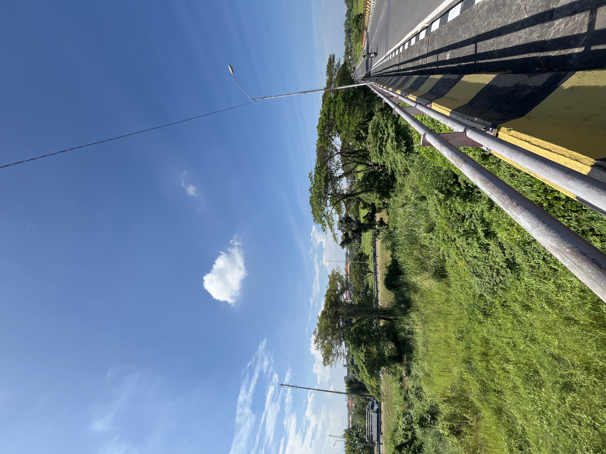 The image captures a clear blue sky with a few wispy white clouds. In the foreground, a bridge railing with yellow and black hazard stripes is visible. The railing is made of metal pipes. Beyond the railing, a road stretches into the distance, with a few motorcycles and a truck driving on it. The road is flanked by lush green vegetation, including trees and tall grass. In the background, there are buildings and more trees, suggesting an urban or semi-urban environment. The scene appears to be a sunny day in Kalisampurno, Indonesia, likely taken from a bridge overlooking a roadside.