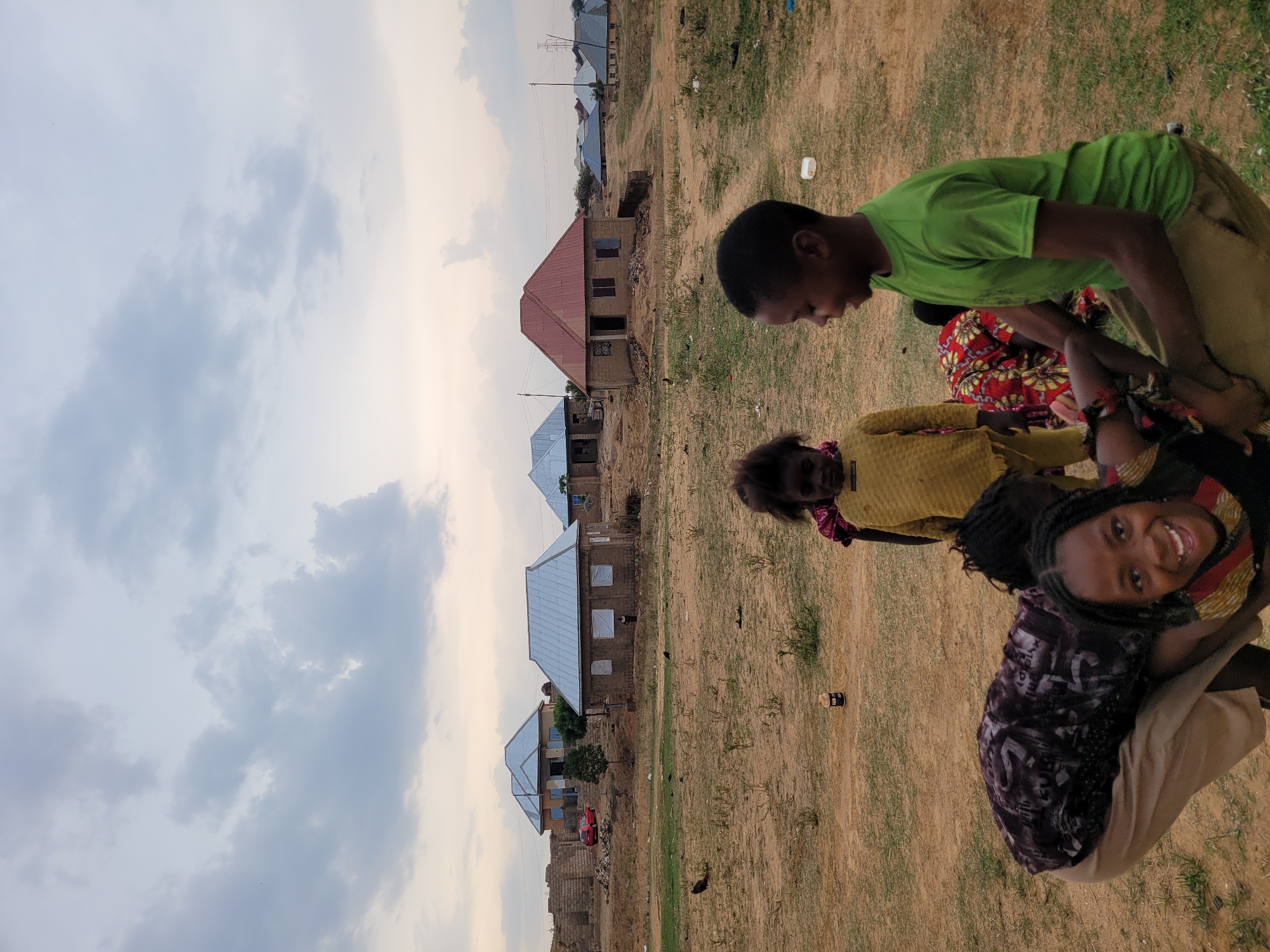 The image depicts a group of people in a grassy, dirt-filled area, seemingly a field or open space. Houses are visible in the background under a cloudy sky. One person, wearing a green t-shirt, is standing and looking down at others in the group. Another person is smiling and looking at the camera. The overall scene suggests a residential area in Barakin Akawo, Nigeria.