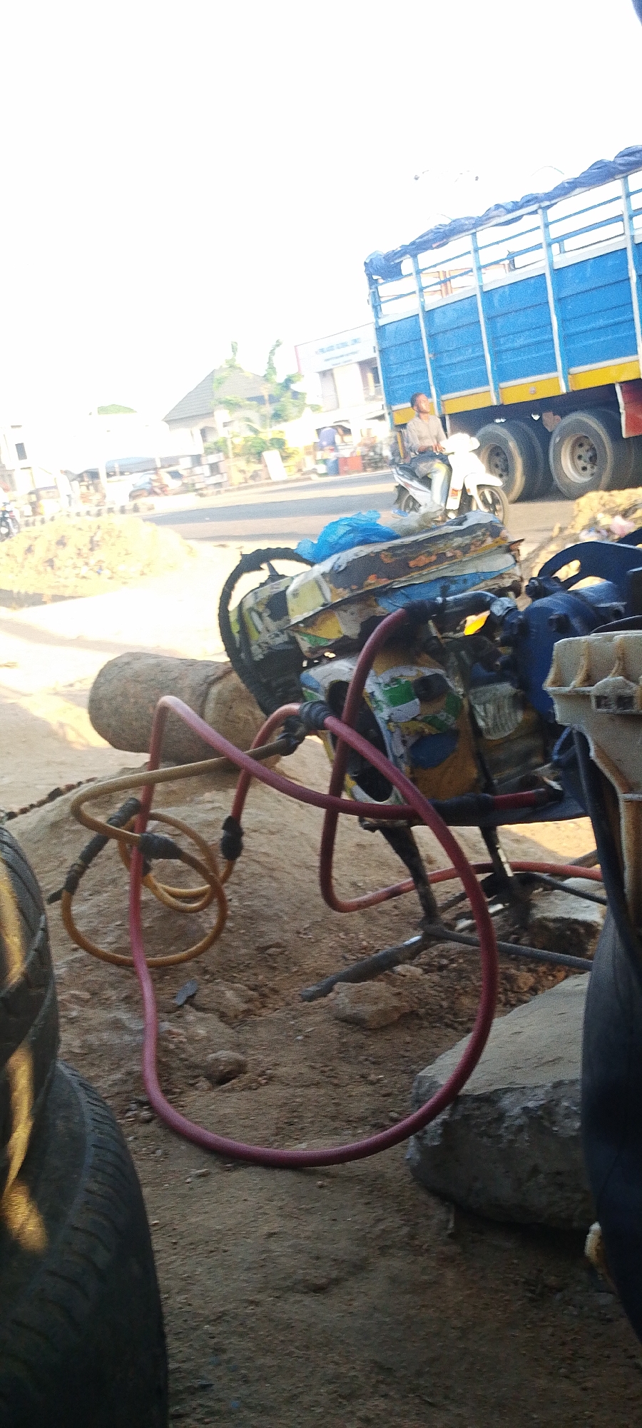 The image depicts an outdoor scene in Lokoja, Nigeria. In the foreground, there is a tire repair setup with various objects including tires, a generator on a stand, and air hoses (one red and one yellow), resting on the ground which is mainly dirt. Behind the generator, there is a street with a blue cargo truck and a man on a motorcycle. There is also a pile of material that appears to be sand. In the background, there are buildings and other signs of a town or city. The scene suggests a roadside tire repair business.