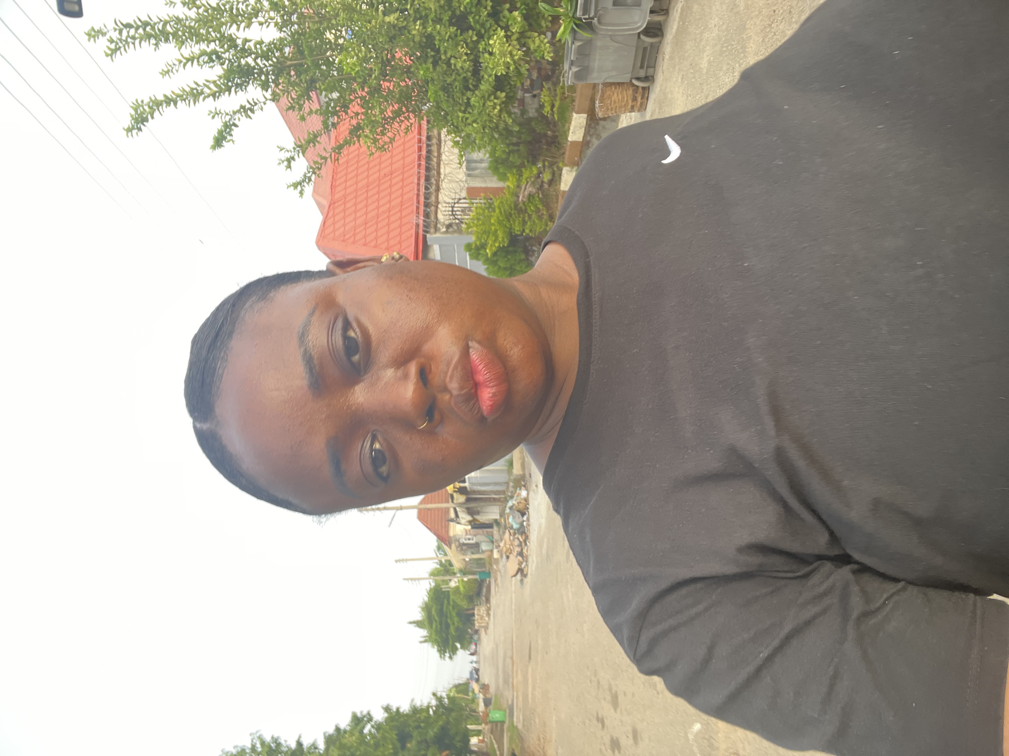 A medium shot depicts an individual with dark skin, wearing a black t-shirt, positioned in an outdoor setting. The background reveals a street scene with buildings featuring red roofs, trees, utility poles, and litter scattered on the pavement. Based on the environment, including the architectural style and street layout, the location is consistent with a residential area in Abuja, Nigeria. The person is facing the camera with a slight upward tilt. The overall impression is a casual snapshot taken outdoors.