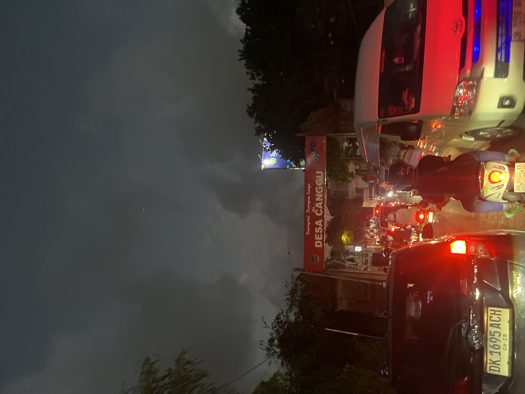 The image depicts a traffic jam at night in North Kuta, Indonesia. A white van with its brake lights illuminated is prominent in the foreground, alongside several motorbikes and other vehicles. The sky is dark and cloudy, suggesting it might be late evening or early night, possibly with impending rain.
A red signpost is visible, with white text that reads "Sampai Jumpa di DESA CANGGU". This translates to "See you in CANGGU VILLAGE", indicating the location. The atmosphere appears to be one of typical urban congestion, with the glow of vehicle lights and streetlights reflecting on the wet or damp road surface.
The scene is filled with vehicles, suggesting a busy thoroughfare. A person is visible riding a motorbike in the lower center of the image. The presence of multiple vehicles and the dark sky contribute to a somewhat muted color palette, with the bright red brake lights and the yellow license plate of the car in the foreground providing pops of color. The overall mood is that of a busy, possibly frustrating, commute.