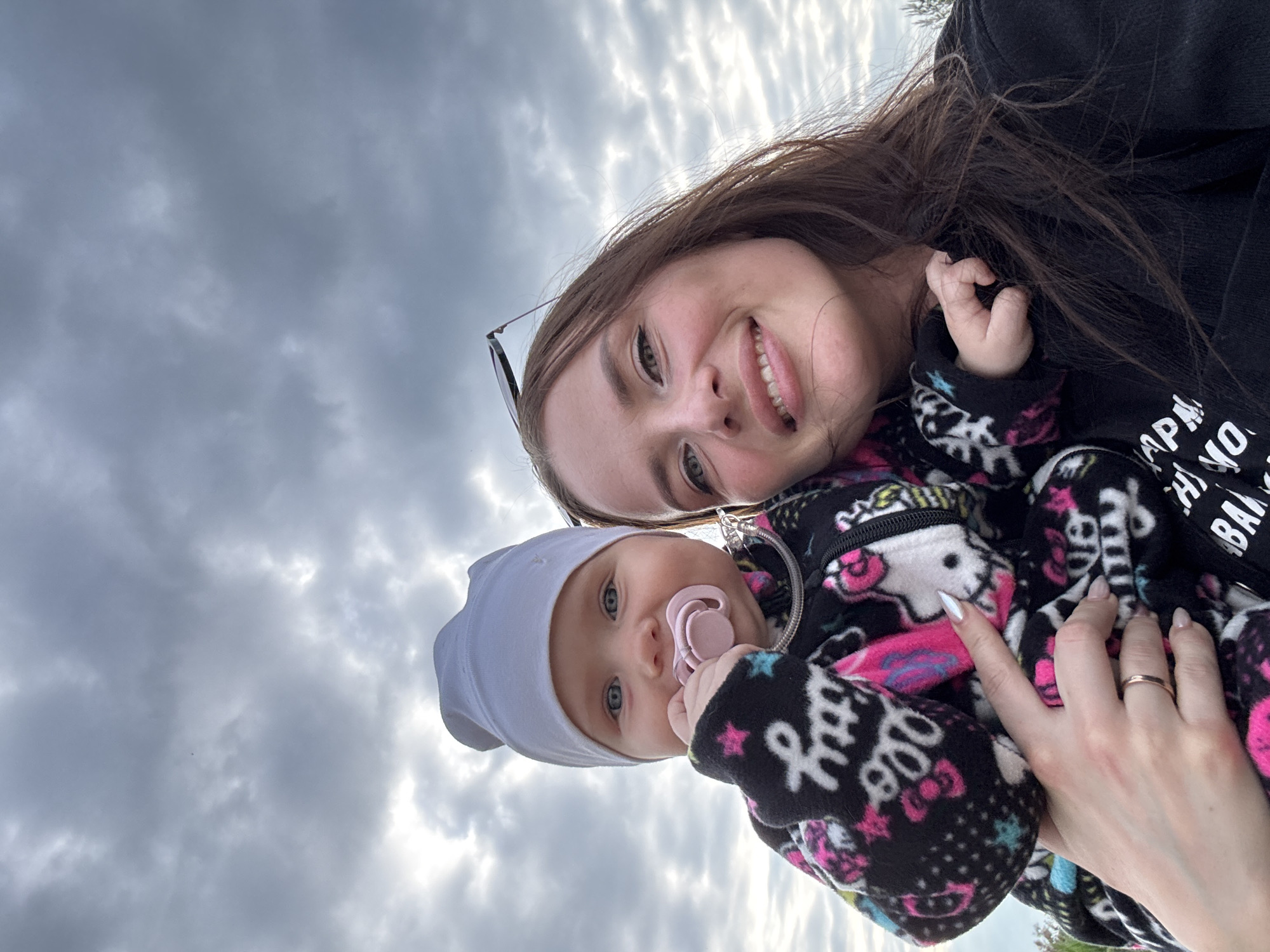 A selfie is taken outdoors with a cloudy sky in the background. A woman and a baby are in the foreground. The woman is on the right side of the frame, looking at the camera with a smile. She has long, brown hair and is wearing a black hoodie with white lettering. Sunglasses are perched on her head. The baby is on the left, held by the woman. The baby is wearing a white beanie and a black, hooded fleece jacket with a Hello Kitty pattern. The baby has a pink pacifier in its mouth and is looking at the camera with wide blue eyes. The woman's left arm is visible holding the baby, and her right arm is partially visible. The woman has a ring on her left ring finger. Part of a dark green bush or tree is visible on the far right of the frame. The scene is a selfie in Novyi, Russia, suggesting an outdoor recreational area or park.