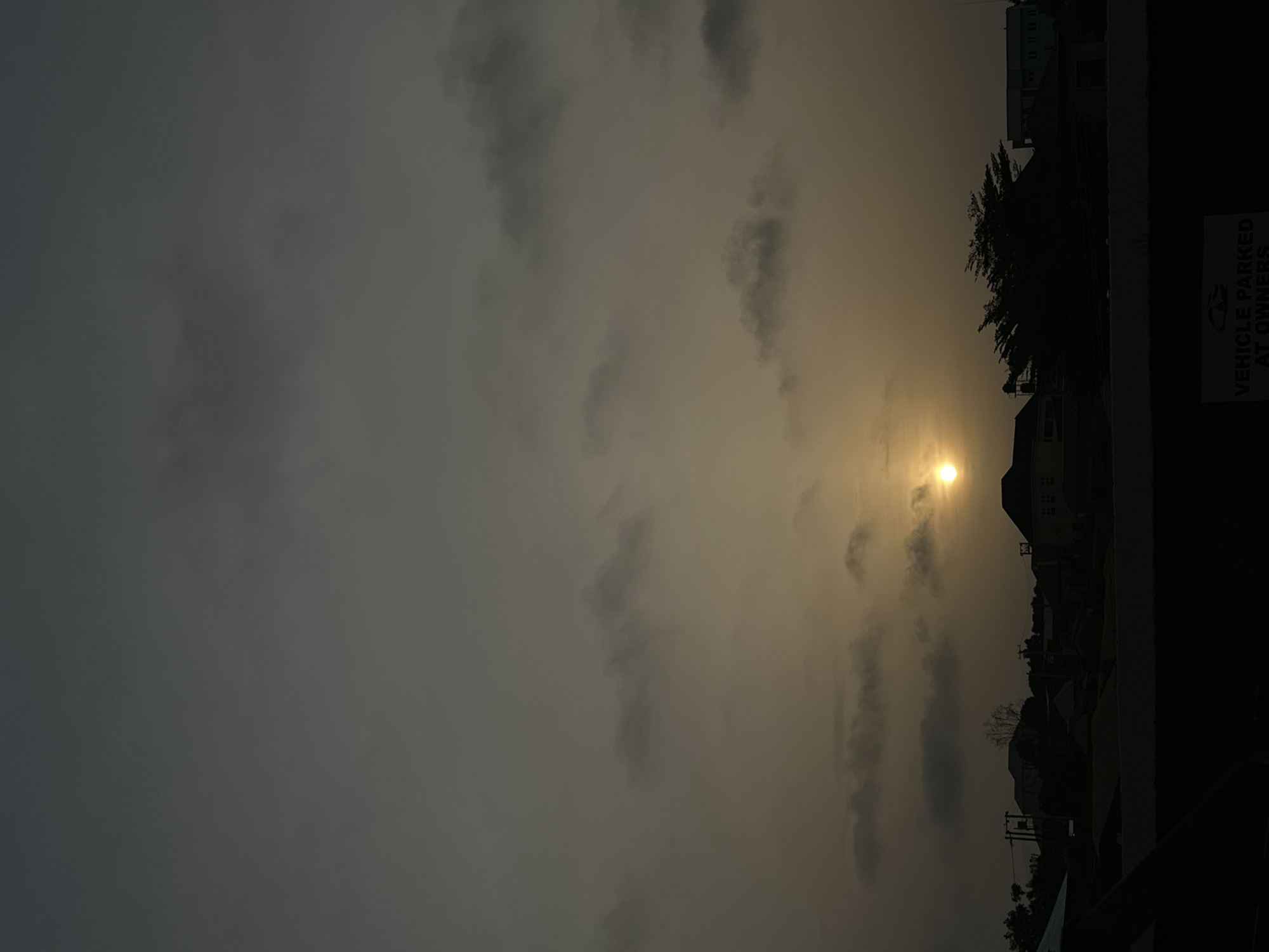 The image captures a view of Jos, Nigeria, at what appears to be either dawn or dusk. The sky is overcast with a layer of grey clouds, with a pale yellow light filtering through, indicating the presence of the sun. The cityscape includes silhouettes of buildings and trees, suggesting a suburban or residential area. A sign reading "VEHICLE PARKED AT OWNERS RISK" is visible on the right side of the frame. The perspective suggests the photo was taken from a slightly elevated position, possibly a building or a balcony.