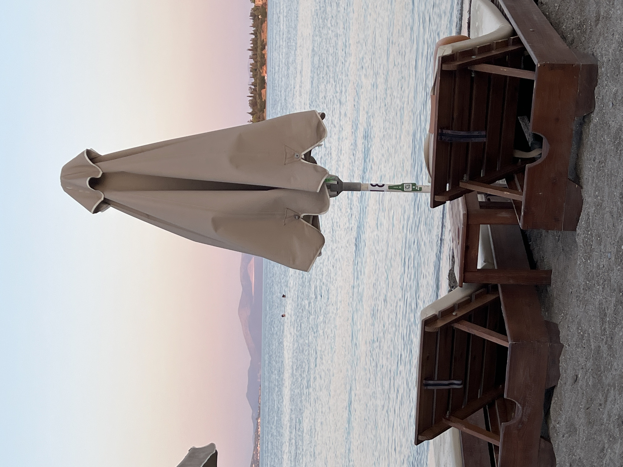 The image depicts a beach scene at dusk in the Amarynthos Municipal Unit, Greece. Two wooden lounge chairs with beige cushions are positioned on the sand, facing the sea. In the center of the frame, a closed, beige beach umbrella stands tall. The calm sea stretches out towards a distant coastline with hills and trees. The sky displays a gradient of soft pink and blue hues. Two figures are visible in the water, appearing as small dark shapes. A small wooden table sits between the lounge chairs. The overall atmosphere is tranquil and serene.