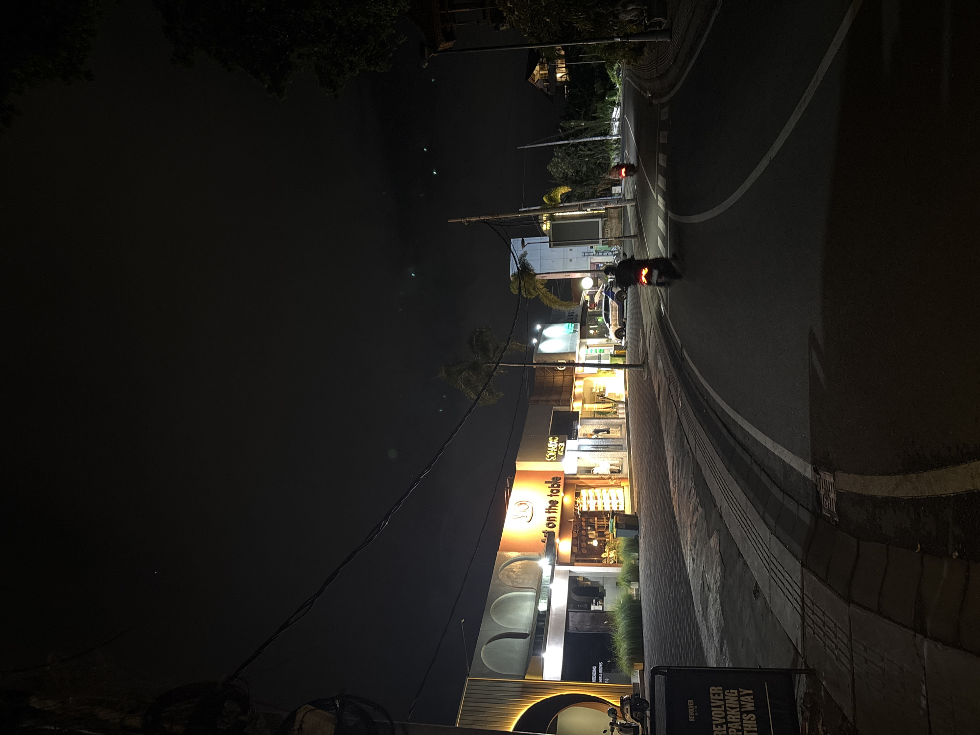 The image depicts a nighttime street scene in North Kuta, Indonesia, characterized by a curved asphalt road flanked by buildings and trees. The lighting is dim, with artificial lights illuminating storefronts and street lamps. A white car with a blue stripe is parked in front of a building, and several motorcycles are visible on the road, with two in motion. One motorcycle in the foreground has its brake lights illuminated. A sign with the text "REVOLVER PARKING THIS WAY" is present on the left side of the street. The sky is dark, with a few faint green lights visible, possibly atmospheric phenomena or distant lights. The architecture of the buildings suggests a commercial or hospitality area, with a mix of modern and traditional elements. The overall atmosphere is quiet and typical of a street at night in a developing urban environment.