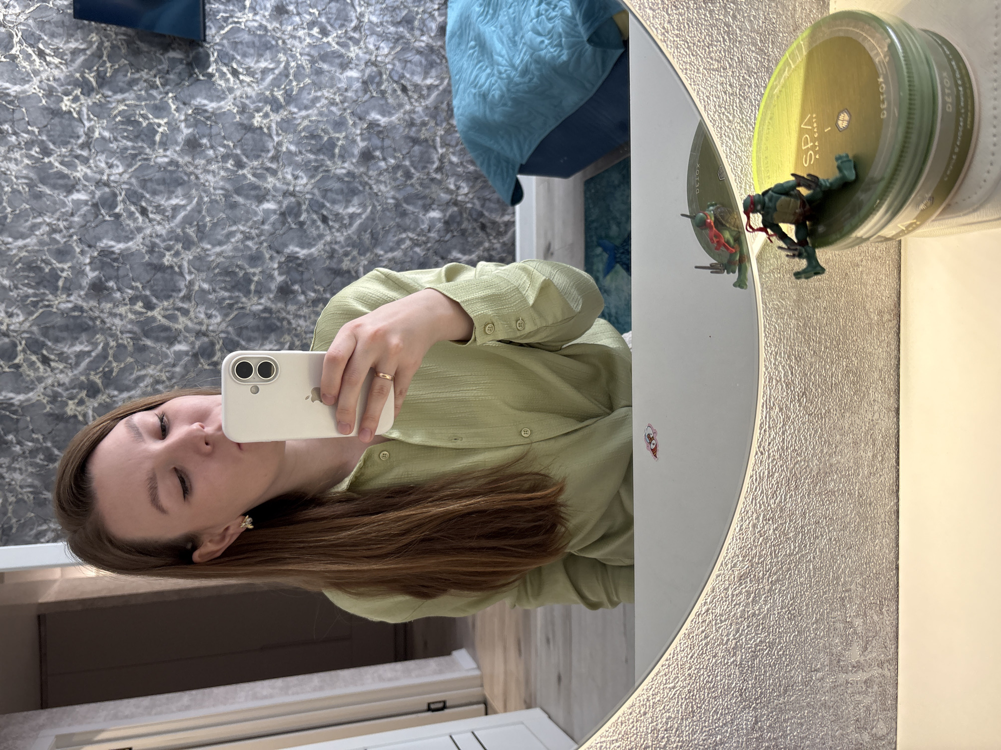 The image shows a person taking a selfie in a mirror. The person has long brown hair and is wearing a light green shirt. The background includes a wall with a grey and white pattern, a glimpse of a blue bed, and a bathroom setting with a vanity. On the vanity, next to the mirror, is a container of "SPA a la carte DETOX" product with a Teenage Mutant Ninja Turtle figure on top. There is no evidence to confirm the location is in Izhevsk, Russia.