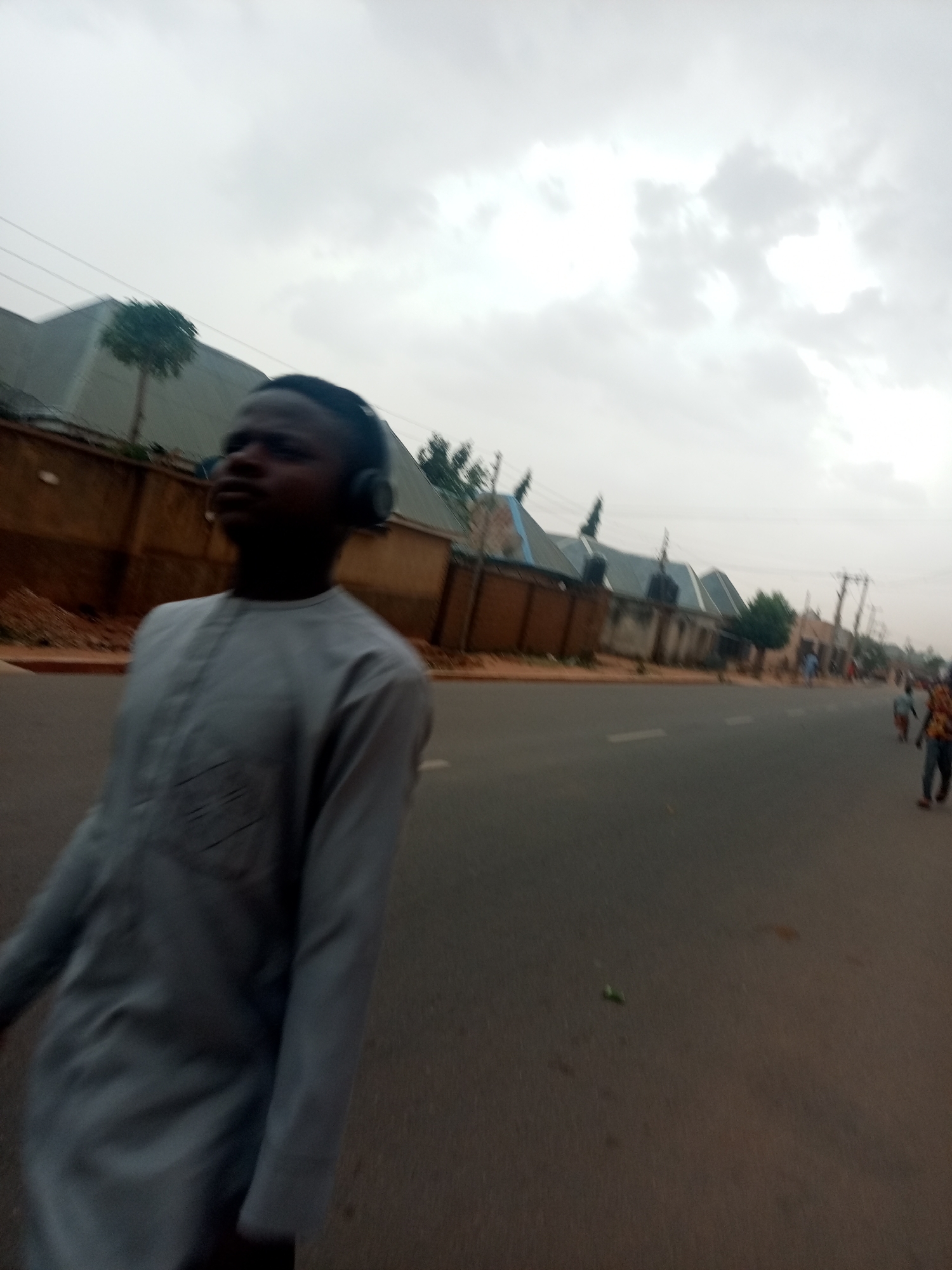 A street in an urban area. A young person wearing headphones and a long-sleeved shirt is walking on the road. Houses and a wall line the street. Other people are walking further down the road. The sky is overcast.