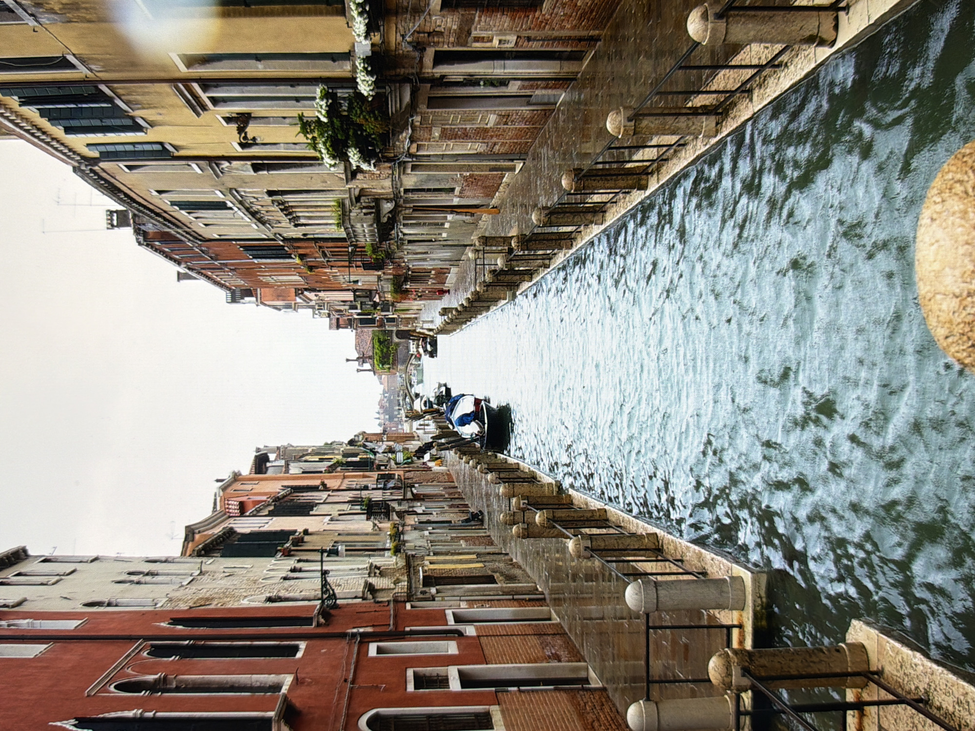 The image depicts a narrow canal in Venice, Italy, with tall, colorful buildings lining both sides. The canal is filled with rippling water, reflecting the overcast sky. A stone walkway runs alongside the canal, bordered by a black metal fence and decorative stone posts. Several gondolas are moored along the walkway, one of which is partially visible in the foreground. The buildings feature traditional Venetian architecture, with arched windows and intricate details. The overall atmosphere is one of quiet, historic charm, characteristic of Venice. The image does not depict any scenes from Kyiv, Ukraine.