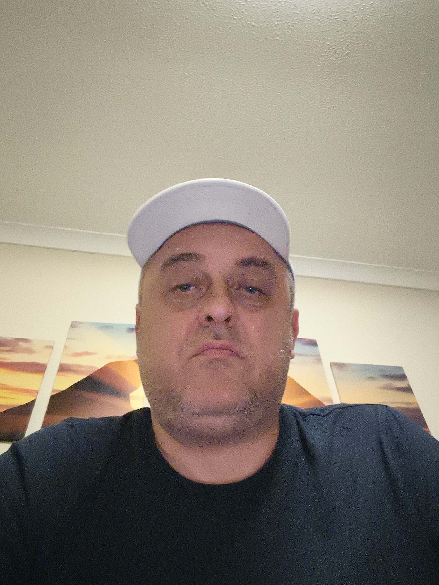 The image displays a close-up, slightly upward-angled portrait of a mid-adult man. He is centered in the frame, shown from the chest up. The man is wearing a light-colored, possibly white or light grey, baseball cap with the brim facing forward. He also wears a dark, plain crew-neck t-shirt. His face features a short stubble or light beard, and his expression is neutral or slightly pensive, with his lips gently pursed. He appears to be looking directly at the camera.
The background consists of a plain, light cream-colored ceiling at the top of the frame, and a wall of the same color behind him. On the wall, several multi-panel art prints are visible, depicting a vibrant sunset or sunrise scene over a landscape, possibly featuring mountains or hills with a warm color palette of oranges, yellows, and blues.
The lighting is consistent with an indoor setting, likely ambient. There are no other visible subjects or objects, and no text is discernible. The overall scene suggests a casual, indoor environment, potentially a home in Huntingdonshire, United Kingdom, with the man posing for a photograph, likely a selfie.