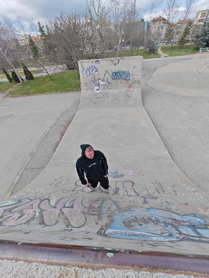 A high-angle, wide-angle photograph depicts a male individual standing on a graffiti-covered concrete skate ramp within Skatepark Shumen, Shumen, Bulgaria. The subject, positioned centrally on the ramp's curved transition, is a male wearing a black hooded sweatshirt with white trim on the sleeves, dark pants, and light-colored athletic shoes. He holds a small, metallic object in his left hand, with his right hand in his pocket, and is looking towards the upper left of the frame. The concrete surfaces of the skate ramp, including its quarter pipe and the curved transition, are extensively covered in multi-colored spray-painted graffiti. Surrounding the main ramp are grey concrete pathways and additional skatepark elements. The background features a park setting with numerous deciduous trees, mostly bare of leaves, and patches of green grass. Multi-story residential buildings are partially visible in the distant background, beyond the trees. The sky is overcast.