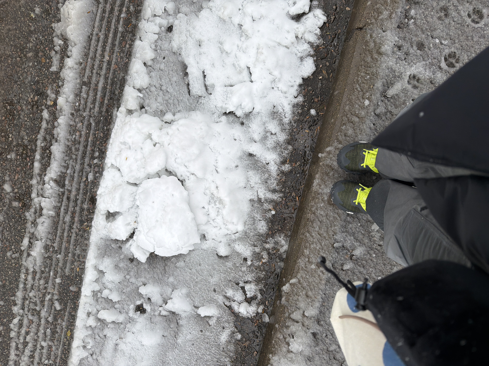An overhead shot captures a person standing on a wintery ground in what appears to be Moscow, Russia. The scene is dominated by a mix of asphalt, snow, and ice, viewed from the waist down, focusing on the person's feet and the immediate surroundings.
On the left side of the image, a dark gray asphalt or paved surface is visible, featuring distinct parallel grooves, likely tire tracks or plow marks, partially covered by a thin, icy layer of snow. This area suggests a pathway or road.
Towards the center and right, the ground transitions to a thicker accumulation of lumpy white snow and slush. Near the person's feet, the snow becomes more granular and wet, exposing patches of dark, wet earth or pavement underneath. Clear paw prints, likely from a dog, are distinctly visible impressed into the slushy ice near the person's right foot.
The person is wearing dark gray pants and a black jacket or coat, with the lower part of the jacket partially obscuring the view on the right. Their feet are clad in robust athletic shoes, featuring dark green or black uppers with bright, neon yellow laces, designed for potentially slippery conditions. A small portion of a light-colored object with blue and brown patterns, possibly a bag or accessory with a black drawstring, is seen in the bottom right corner.
The weather conditions are cold and overcast, with melting snow and ice suggesting temperatures around freezing. The overall impression is of a typical winter day outdoors, with the person likely walking or pausing on a snow-covered path. There is no visible text in the image.