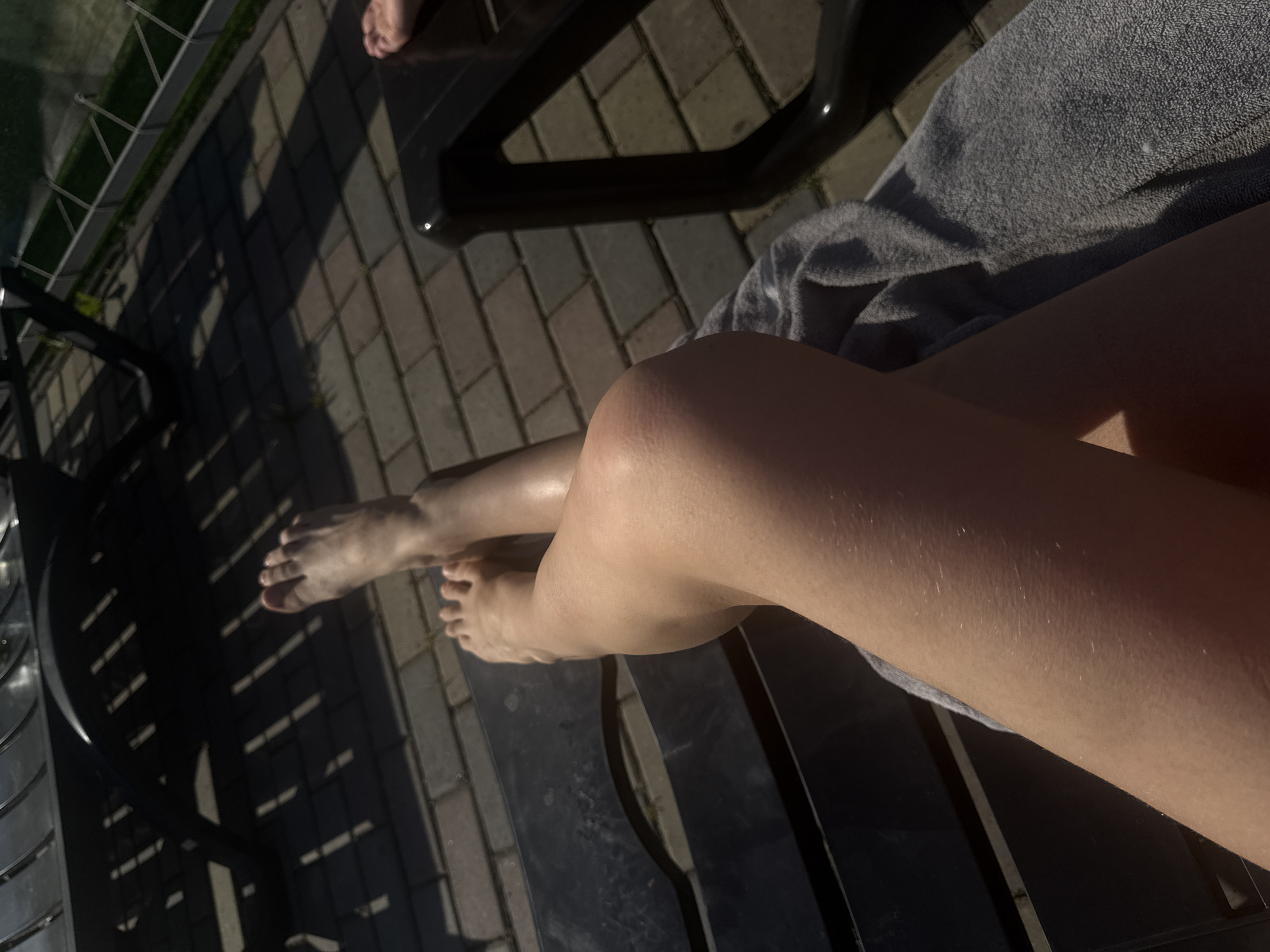 The image depicts a scene outdoors, likely near a pool or patio area given the paving stones, lounge chair, and partial view of what appears to be a fence near greenery. In the foreground are a pair of light-skinned human legs crossed at the knees, resting on a black lounge chair. A grey towel is draped under the thighs. The person's bare feet are visible at the edge of the chair. Another person's bare foot is partially visible in the top center of the frame. The scene appears to be bathed in sunlight, creating sharp shadows from the furniture. The exact location in Гамово, Russia cannot be determined from the image alone.