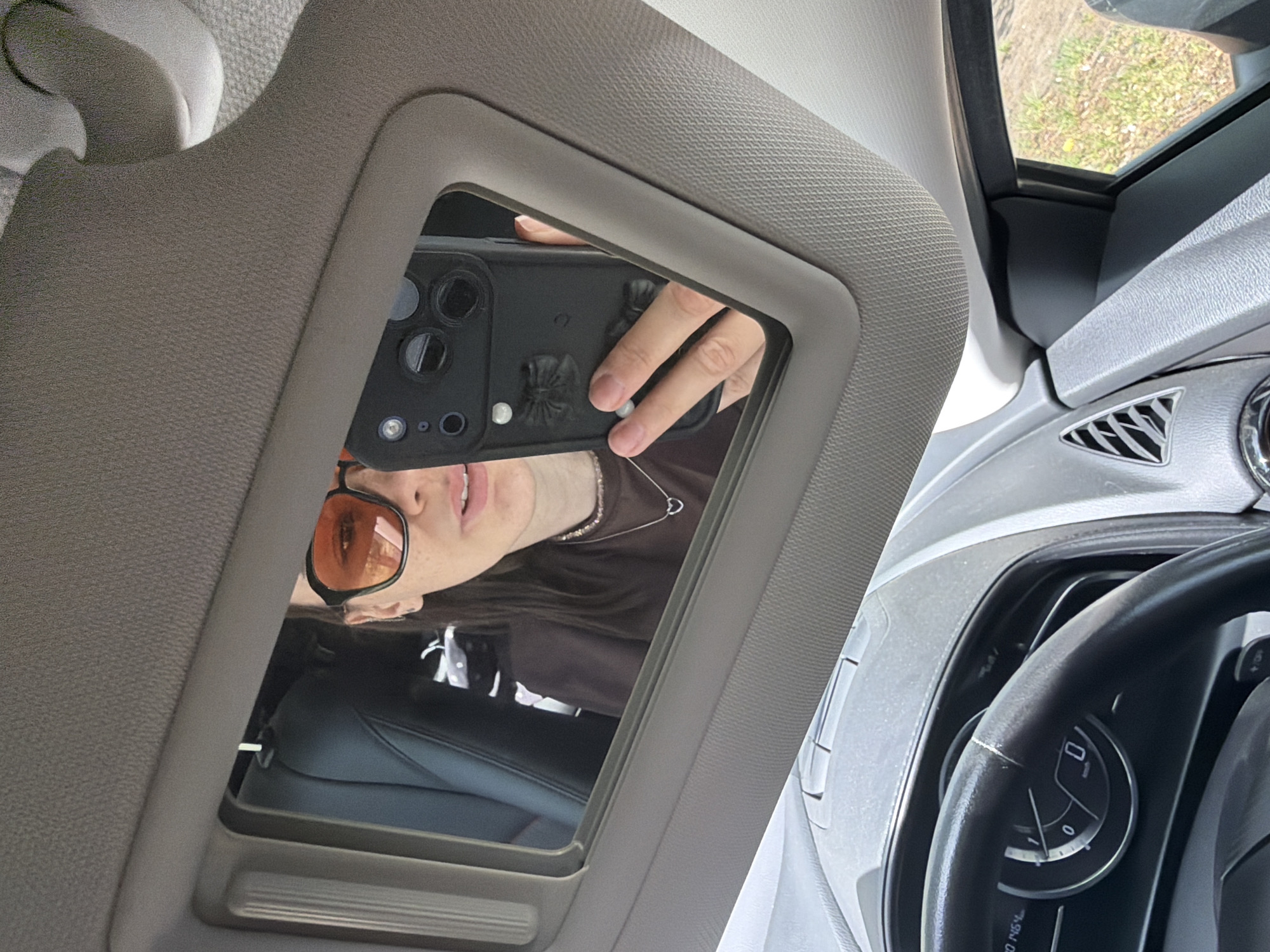 The image depicts a selfie taken inside a car, reflected in a visor mirror. A person with orange-tinted glasses, a brown shirt, and a silver necklace with a heart pendant is visible in the mirror's reflection, holding a black phone with decorations on its case. The car's dashboard, steering wheel, and front seat are partially visible, suggesting the interior of a passenger vehicle. The scene suggests the image was taken inside a car, but there is no information provided to support the location being in the city of Chaikovsky, Russia.