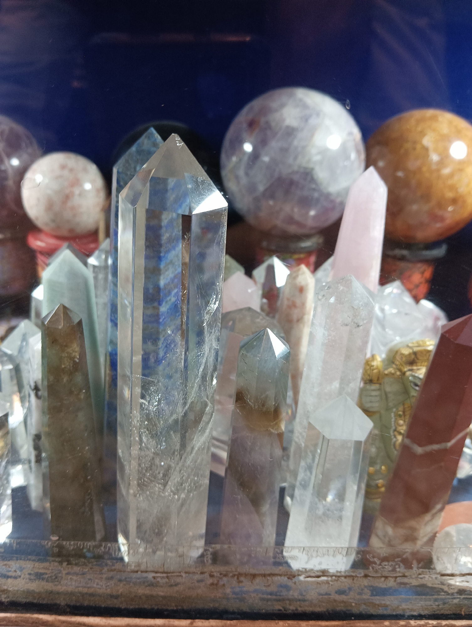 This media file showcases a close-up, eye-level view of a collection of assorted crystals, predominantly clear quartz and some other specimens with blue and pinkish hues. The crystals are arranged in a shallow display, possibly on a shelf or within a cabinet. The background is a dark, solid blue, creating a stark contrast with the light-colored crystals.
The subjects are primarily the crystals themselves, which vary in size, shape, and clarity. Some appear as sharp, elongated points, while others are more rounded, spherical, or faceted. One prominent crystal in the foreground is a tall, clear quartz point with internal fractures and inclusions, revealing blue-banded patterns within. Other noticeable crystals include a pale pink point, a smoky-colored point, and a reddish-brown point. The arrangement suggests a decorative or collectible display rather than an active event.
Notable details include the lighting, which appears to be ambient and possibly artificial, highlighting the reflective surfaces of the crystals. There are no visible people, text, or clear indicators of time of day or weather. The setting is indoors, suggested by the controlled lighting and background. The overall impression is one of stillness and appreciation for the natural beauty and forms of the crystals. The presence of some dust or debris on the display surface in the foreground might suggest the location is a shop, a personal collection, or a spiritual or healing space. Given the location context of Vashisht, India, a spiritual or therapeutic setting is plausible.