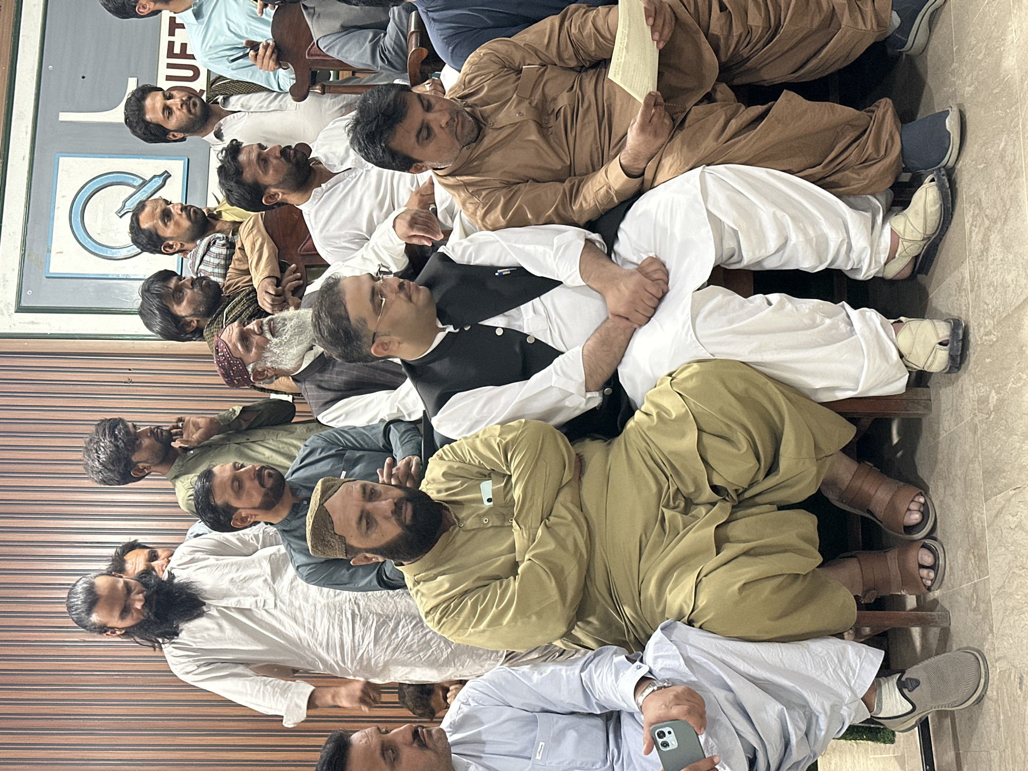 A group of approximately 14 adult males are seated close together on wooden chairs in what appears to be an indoor gathering in Quetta City Tehsil, Pakistan. They are diverse in age, with some having beards of varying lengths and colors. The men are dressed in traditional Pakistani clothing, including shalwar kameez in different colors such as white, brown, olive, and light blue. One man wears a black vest over his white clothing.
Some individuals are looking up or around the room, while others appear to be looking down or at each other. One man is reading from a piece of paper. A mobile phone is visible in the hand of one man. A sign with blue and white lettering is visible on a wall. The floor is tiled. The setting suggests a casual meeting or gathering.