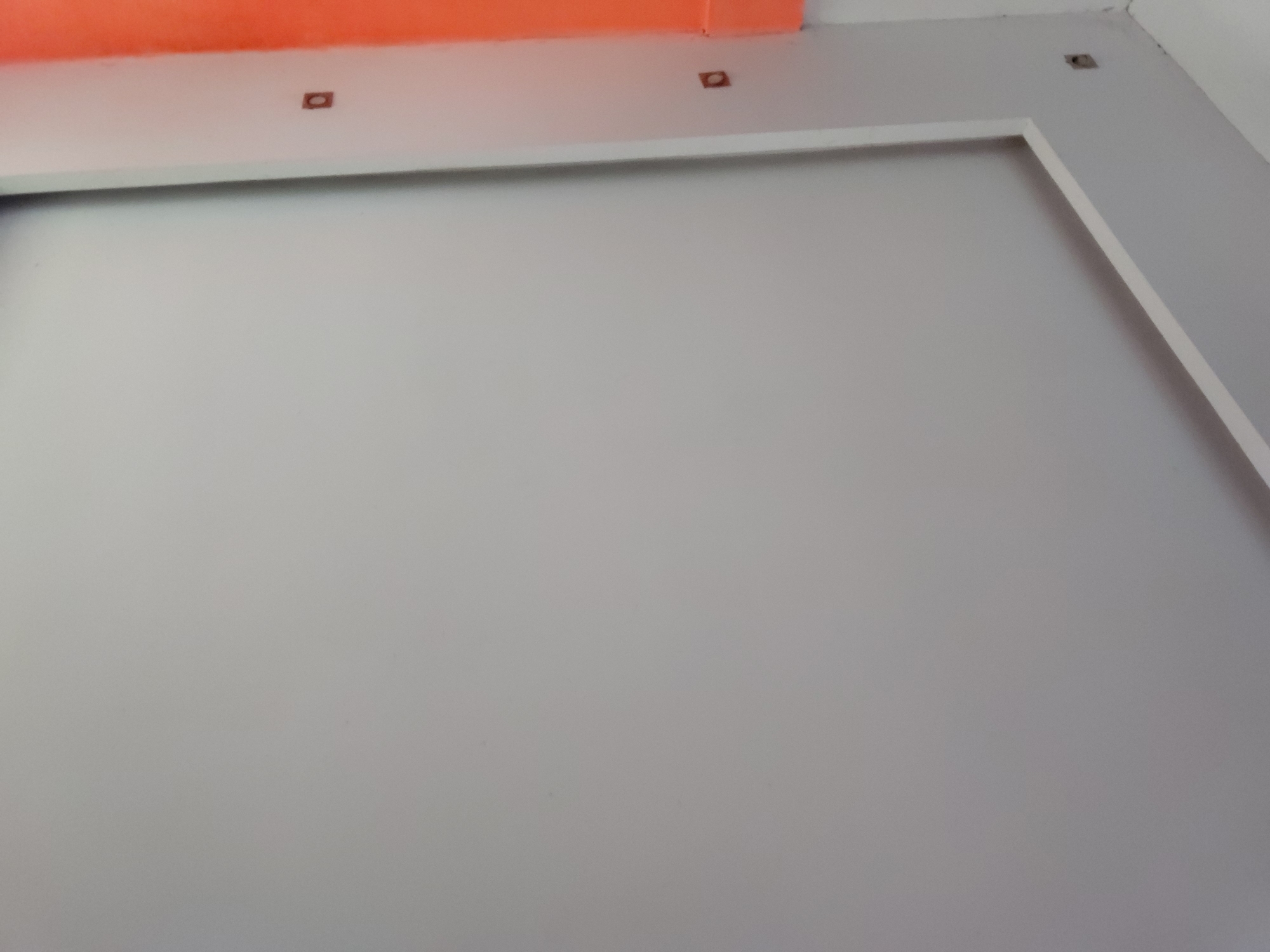 The image captures an interior architectural detail, primarily focusing on a large, plain light gray wall. This wall, which occupies the majority of the frame, is smooth and evenly lit, suggesting an indoor setting. Along the top right section of the gray wall, there is a distinct white rectangular frame, outlining what appears to be a recessed area or a decorative panel.
Above this framed section and the upper part of the gray wall, a horizontal band of white separates it from a vibrant orange-red element at the very top edge of the image. This orange-red component could be a ceiling beam, a soffit, or a painted upper wall section, providing a strong color contrast to the neutral tones below. Embedded within the white band, just below the orange-red section, are three small, dark, rectangular openings or cutouts. Two are visible towards the center-left, and a third is further to the right, near the corner where the white frame meets the ceiling line. These openings may be unfinished electrical points, fixture mounts, or ventilation details.
The scene is static, devoid of any people or discernible activity. The lighting is uniform, characteristic of an indoor environment, possibly illuminated by artificial light or indirect natural light. No text is visible anywhere in the image. While the location context is provided as Santa Ana, Venezuela, the visual elements themselves offer no specific geographical cues, presenting a generic modern interior aesthetic.
