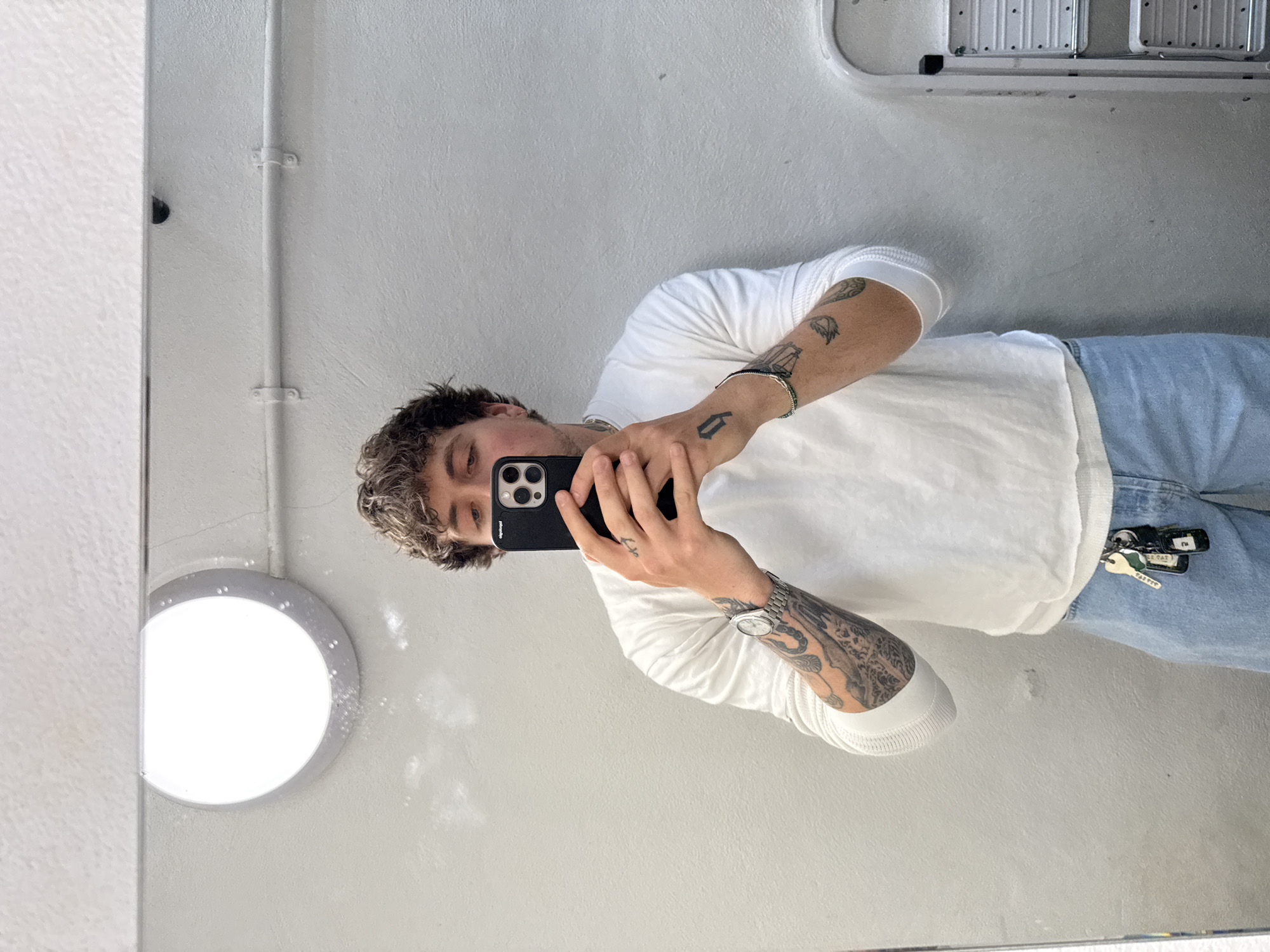 A person with curly brown hair is holding a black iPhone in front of their face, taking a selfie in a mirror. The person is wearing a white long-sleeved shirt and light blue jeans with keys hanging out of the back pocket. The background features a white wall with a circular light fixture and a metal pipe running vertically along the wall. The environment appears to be an indoor space, possibly a bathroom or utility room. The location in the city of London, United Kingdom cannot be determined.