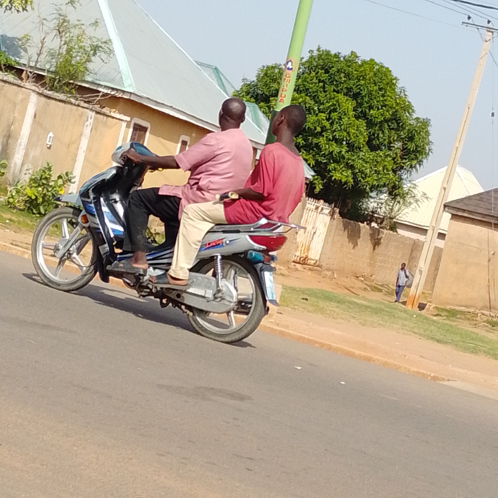 Two individuals are riding on a motorcycle on a paved road in Tempure, Nigeria. The rider in front is wearing a pink shirt and black pants, while the passenger behind is wearing a red shirt and light-colored pants. The motorcycle is silver and blue. Behind them, a street light pole with some writing on it is visible, along with trees, houses with light-colored roofs, and a brick wall. Further in the background, a person in a light blue shirt and dark pants is walking, carrying a bag. The sky is clear and blue.