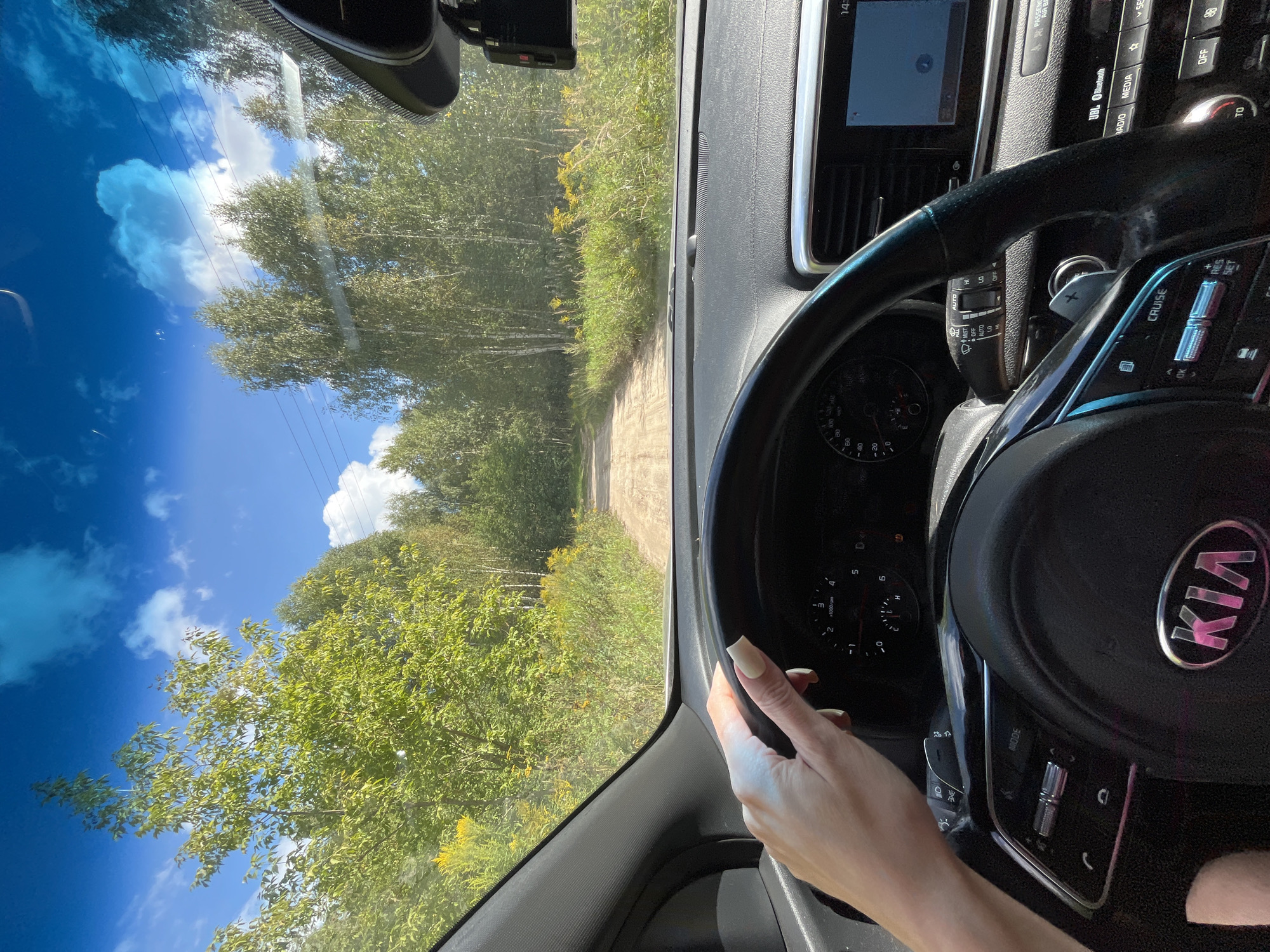 An interior shot of a car shows a person's left hand with long, manicured nails resting on the steering wheel. The steering wheel has various buttons and controls, including the "Kia" logo. The dashboard displays a speedometer and tachometer, and a touch screen is visible to the right. Through the windshield, a dirt road winds through a landscape of green trees and foliage under a bright blue sky with scattered white clouds. Power lines are visible in the distance. The scene is consistent with a drive on a rural road in the outskirts of a city like Nagorny, Russia, during daylight hours.