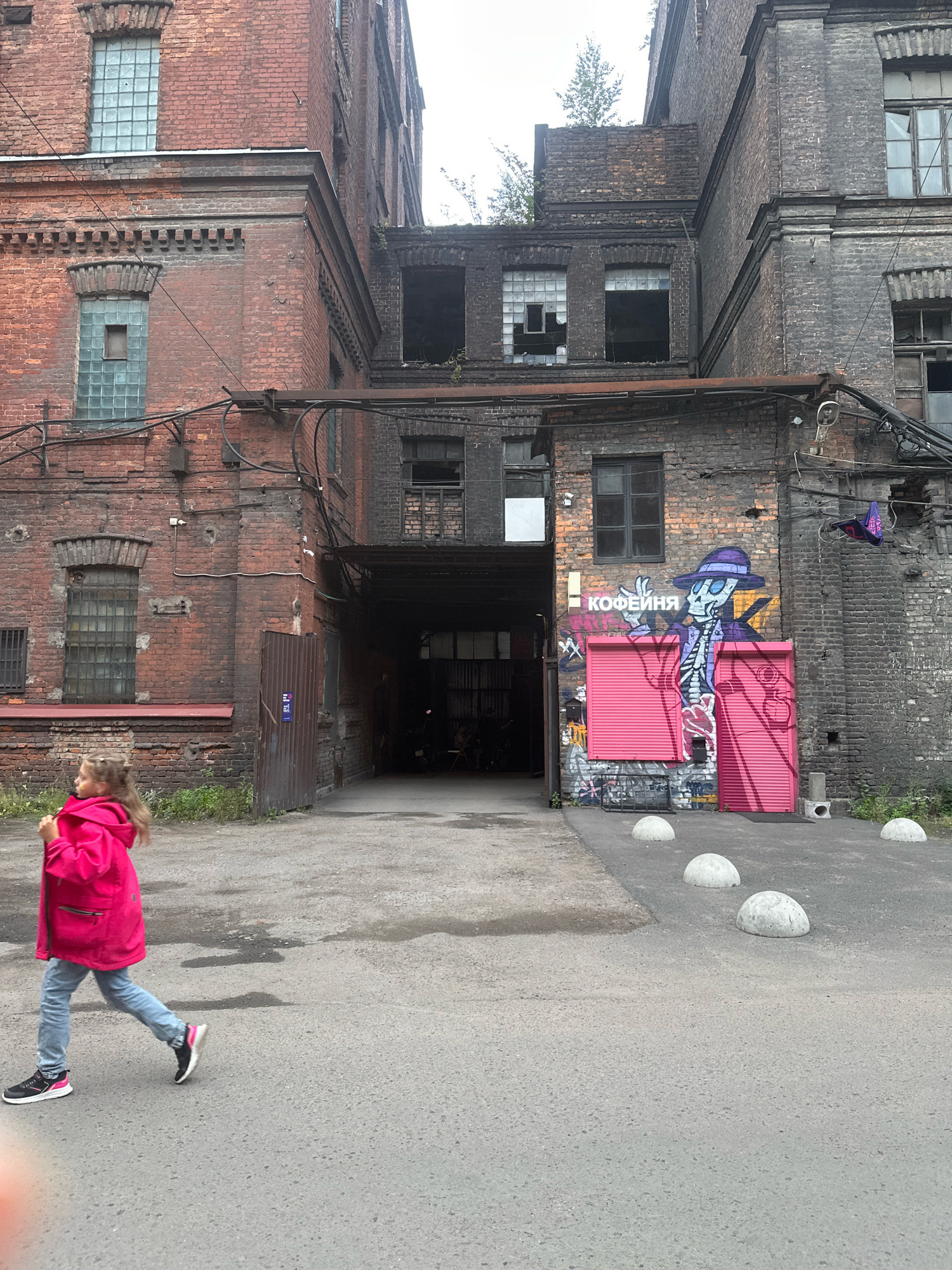 The image depicts a street scene in Saint Petersburg, Russia, featuring an old brick industrial building with a dark, weathered facade and several windows, some of which are broken. A young girl in a bright pink jacket and blue jeans is walking on the asphalted ground towards the left of the frame. Graffiti adorns the brick wall, including a skeletal figure in a hat and a sign that reads "КОФЕЙНЯ," which translates to "coffee shop." Several rounded concrete bollards are placed along the street. The sky is overcast. The building appears to be a former factory or warehouse, characteristic of some industrial areas in Saint Petersburg.