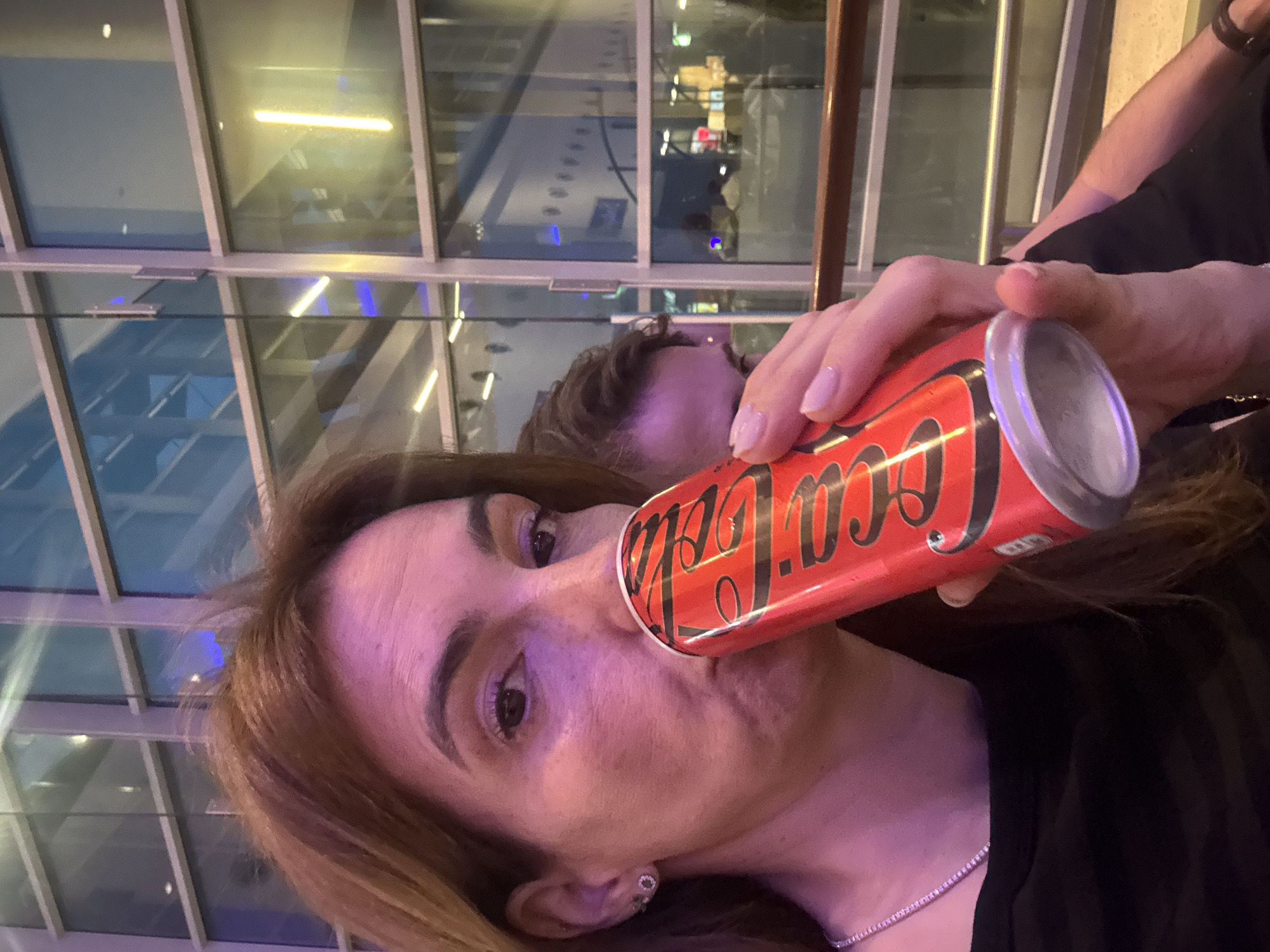 A close-up, selfie-style photograph captures a woman taking a drink from a red Coca-Cola can in an indoor, modern public space during the evening or night. The woman, with light brown hair and dark eyes, is centered in the frame, looking directly towards the camera. She wears a dark top, a delicate silver necklace, and a small, light-colored earring. Her right hand (from the viewer's perspective) holds the can to her mouth. Her fingernails are painted a subtle, light hue.
The can is prominently displayed, featuring the iconic black "Coca-Cola" script against its vibrant red background. The ambient lighting in the scene, likely artificial, casts a distinct purple-pink glow across the subjects and the environment, particularly noticeable on the woman's face and hair.
The background is dominated by a large glass and metal architectural structure, possibly a wall or ceiling, which reflects numerous artificial lights from within the building. These reflections create blurred patterns that hint at other areas of the facility, such as shops or corridors. The top of another person's head and shoulder are faintly visible in the out-of-focus background. The modern glass and steel elements suggest a public venue like an airport terminal, a shopping mall, or a contemporary building in Vila Nova de Gaia, Portugal. The woman's expression is neutral and relaxed.