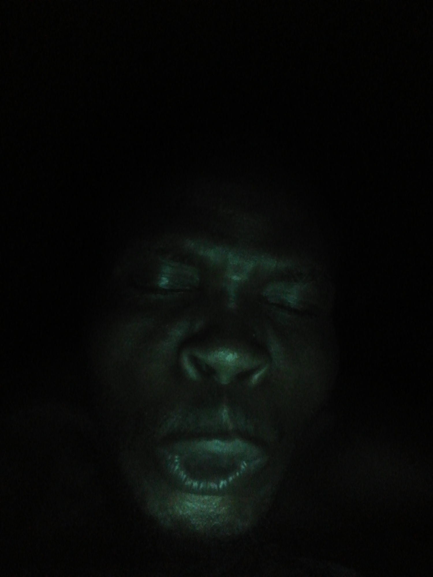 A close-up, darkly lit image reveals a person's face. The individual has their eyes closed and mouth slightly open. The overall coloration of the image has a greenish tint. The environment is overwhelmingly dark, obscuring any clear background details. The location and city cannot be identified due to the lack of contextual elements.