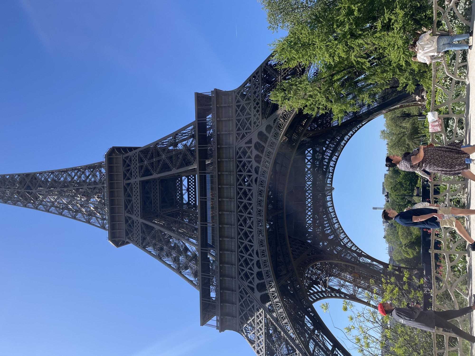 This photograph captures a striking low-angle view of the Eiffel Tower in Paris, France, on a clear, sunny day. The magnificent iron lattice structure of the tower dominates the left side of the frame, extending upwards towards a cloudless, vibrant blue sky. Its intricate engineering and design are clearly visible from this perspective.
On the right side of the image, several people are seen in various positions, appearing to be tourists enjoying the landmark. A decorative metal railing, possibly part of a viewing platform or pathway, separates these individuals from a lower area. Among the visible figures are a person in a dark patterned dress, another in a white top and jeans, a man in a dark blue shirt and light shorts, and a person wearing a dark top and a red cap. Lush green trees and foliage frame the scene on the right, adding a natural element to the urban landscape.
The weather is bright and clear, indicative of a pleasant day for sightseeing. A faint airplane contrail is visible high in the expansive blue sky. The time of day appears to be late morning or early afternoon, given the strong, natural illumination.
A notable detail on the structure of the Eiffel Tower itself is the presence of inscribed names on a horizontal beam on the first level. These are the names of 72 French scientists, engineers, and mathematicians, with some legible names including "DUMAS," "FOURNIER," "CARRON," "SAGUET," and "BIERAT." The overall scene portrays a typical tourist experience at one of the world's most famous monuments.