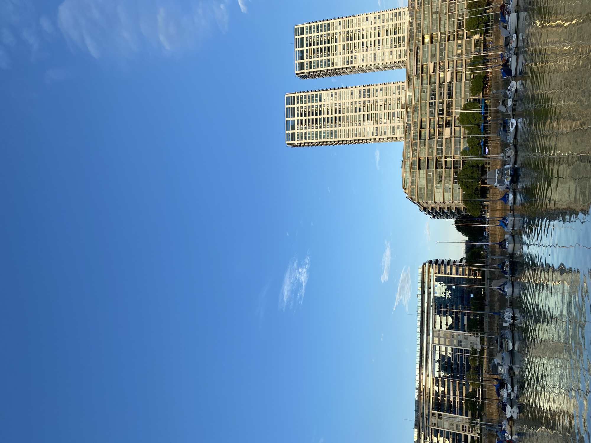 The image shows a harbor in Buenos Aires, Argentina, with modern buildings reflected in the water. Two tall, rectangular buildings stand side-by-side, and another connected building extends to the right. A row of boats is docked along the harbor, with their masts visible. The sky is clear and blue. The water's surface is rippled, distorting the reflections.
