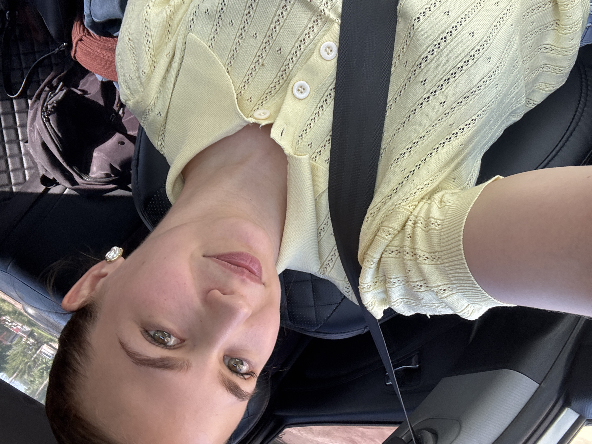 A young adult female, with light skin, green eyes, and light brown hair pulled back, is depicted in a selfie taken inside a vehicle. She wears a light yellow, short-sleeved knitted top with a collar and three visible white buttons, featuring an openwork pattern. A dark seatbelt crosses her chest. A prominent, large, square-cut light-colored earring in a gold-toned setting is visible on her right ear. Her expression is a slight smile. The vehicle's interior features a black quilted seat and dark trim. In the background, behind the subject, a dark backpack, another dark bag, a rust-colored knitted garment, and a blue garment are present on what appears to be the rear seat or floor. Through the window, a blurred outdoor scene shows green trees and a light-colored building, consistent with a suburban or residential setting. No definitive visual markers confirm the location as Chaikovsky, Russia.