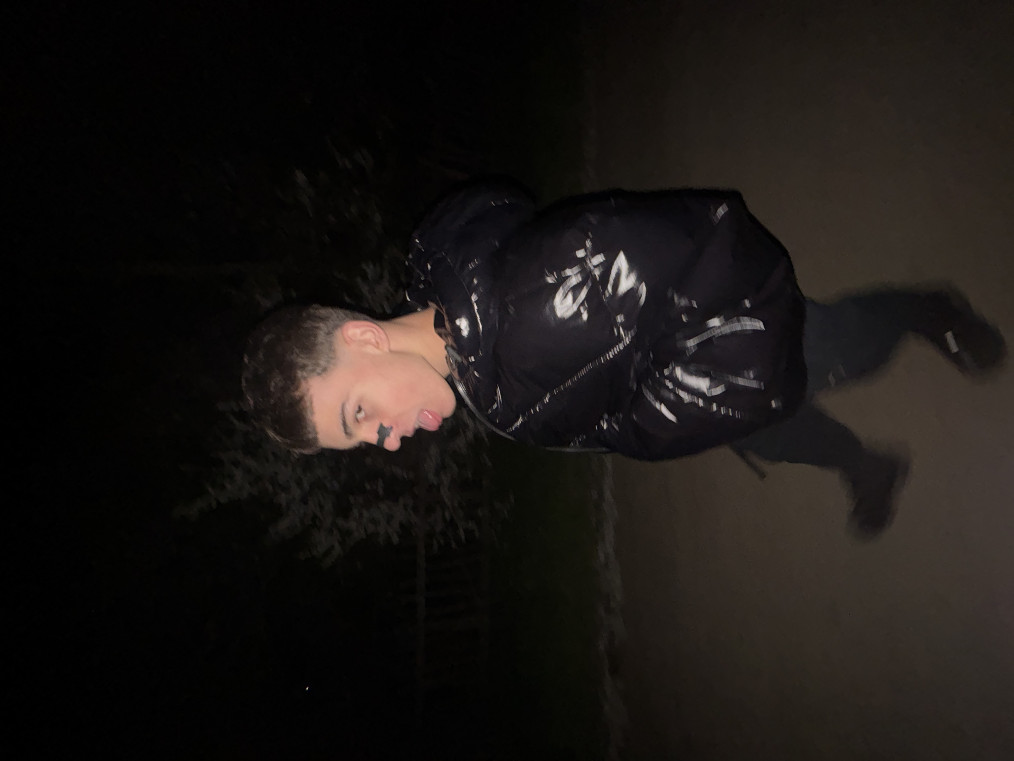 A young man stands on a paved surface outdoors at night. He wears a dark, glossy puffer jacket and dark pants. The background is dark, with some vegetation visible on the left and a wall on the right. The lighting is poor.