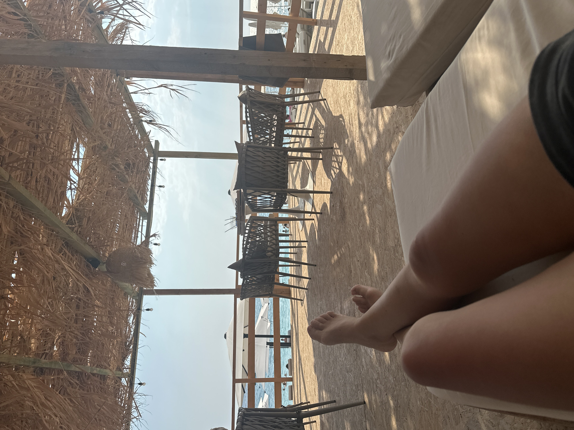 The image shows a first-person perspective of someone relaxing on a beach lounger under a thatched roof structure. In the foreground, the legs and feet of the person are visible, with their legs crossed. Behind them, the scene opens up to reveal a beachside setting. Several woven chairs and tables are arranged on a wooden deck. Beyond the deck, there is a wooden railing overlooking the ocean. The sea is a clear blue, with some white lounge chairs and umbrellas visible on the sandy beach. A few people are seen in the water and on the beach in the background. The sky is bright with some scattered clouds. The overall ambiance is one of leisure and relaxation in a coastal environment.