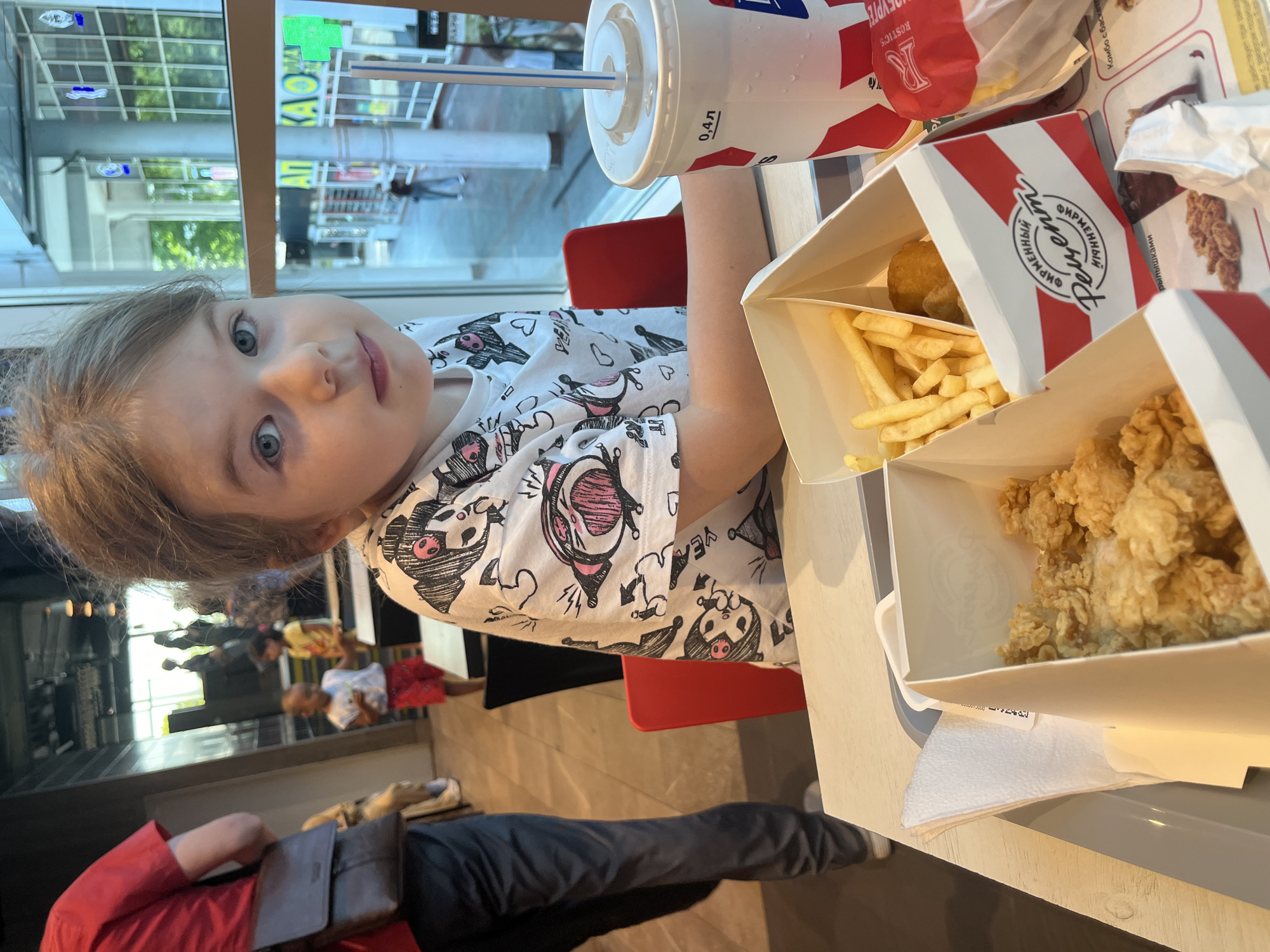 The image shows a young girl with blonde hair and blue eyes sitting at a table in what appears to be a fast-food restaurant, likely KFC, given the branding on the food containers and drink cup. She is wearing a white t-shirt with a playful pattern of cartoon pigs and hearts. In front of her are several boxes of fried chicken and french fries, along with a large cup of soda. A straw is sticking out of the cup. The girl is looking directly at the camera with a neutral expression.
In the background, through a large window, we can see reflections of people and a street scene, suggesting an urban setting. The lighting indicates it is daytime. The overall mood is casual and relaxed, as one would expect in a fast-food eatery. Visible text includes "0,4 л" on the cup, and branding such as "R ROSTIC'S" and "ФИРМЕННЫЙ Рецепт" (Firm Recipe) on the food packaging, which is in Russian. The location context is identified as Sochi, Russia. Further in the background, other patrons can be seen seated and walking. The bottom portion of the image shows a person in a red shirt and dark pants, and another person's legs in dark pants.